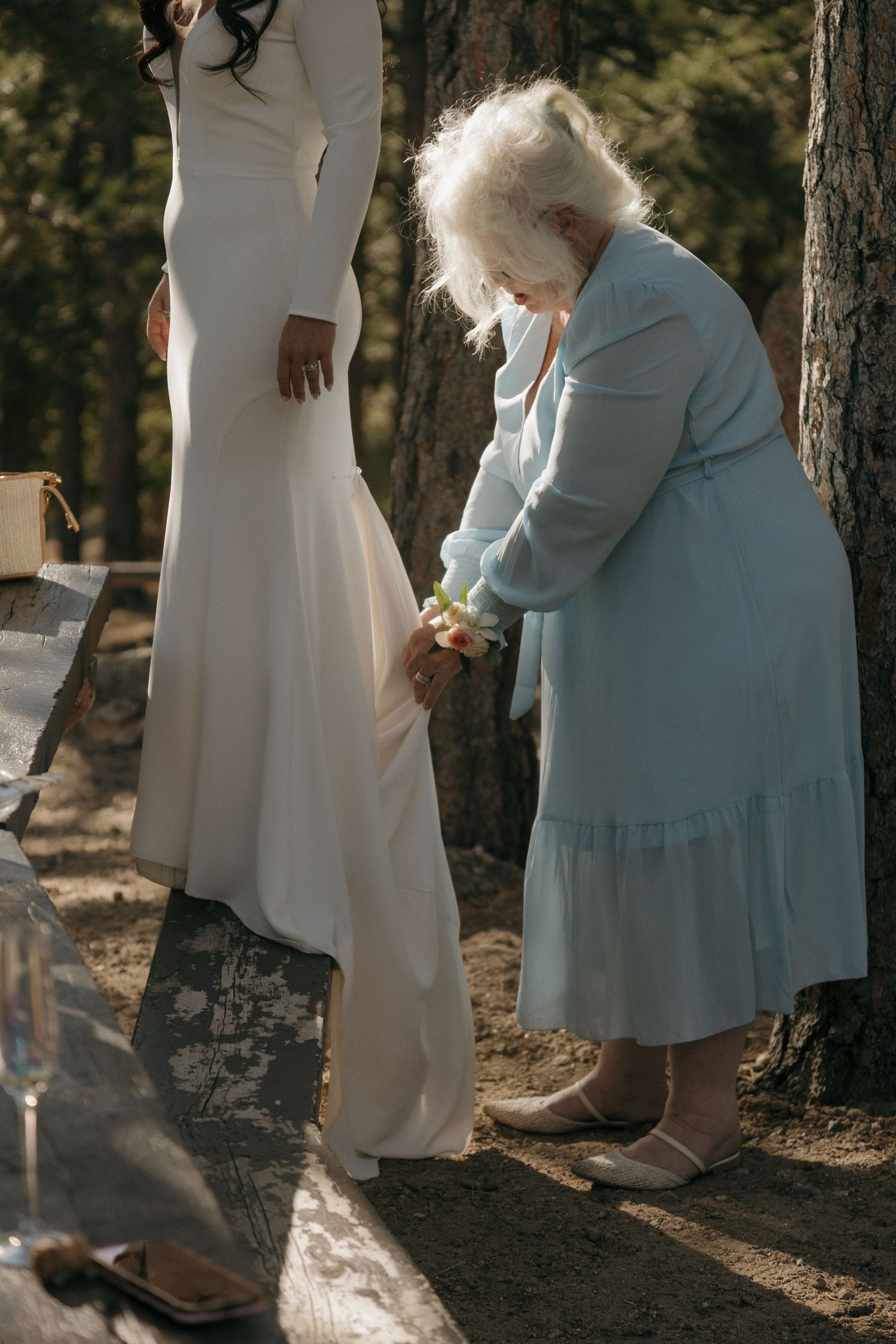 Elderly woman in blue dress holding hands with bride in white long-sleeve wedding gown in sunlit forest setting.