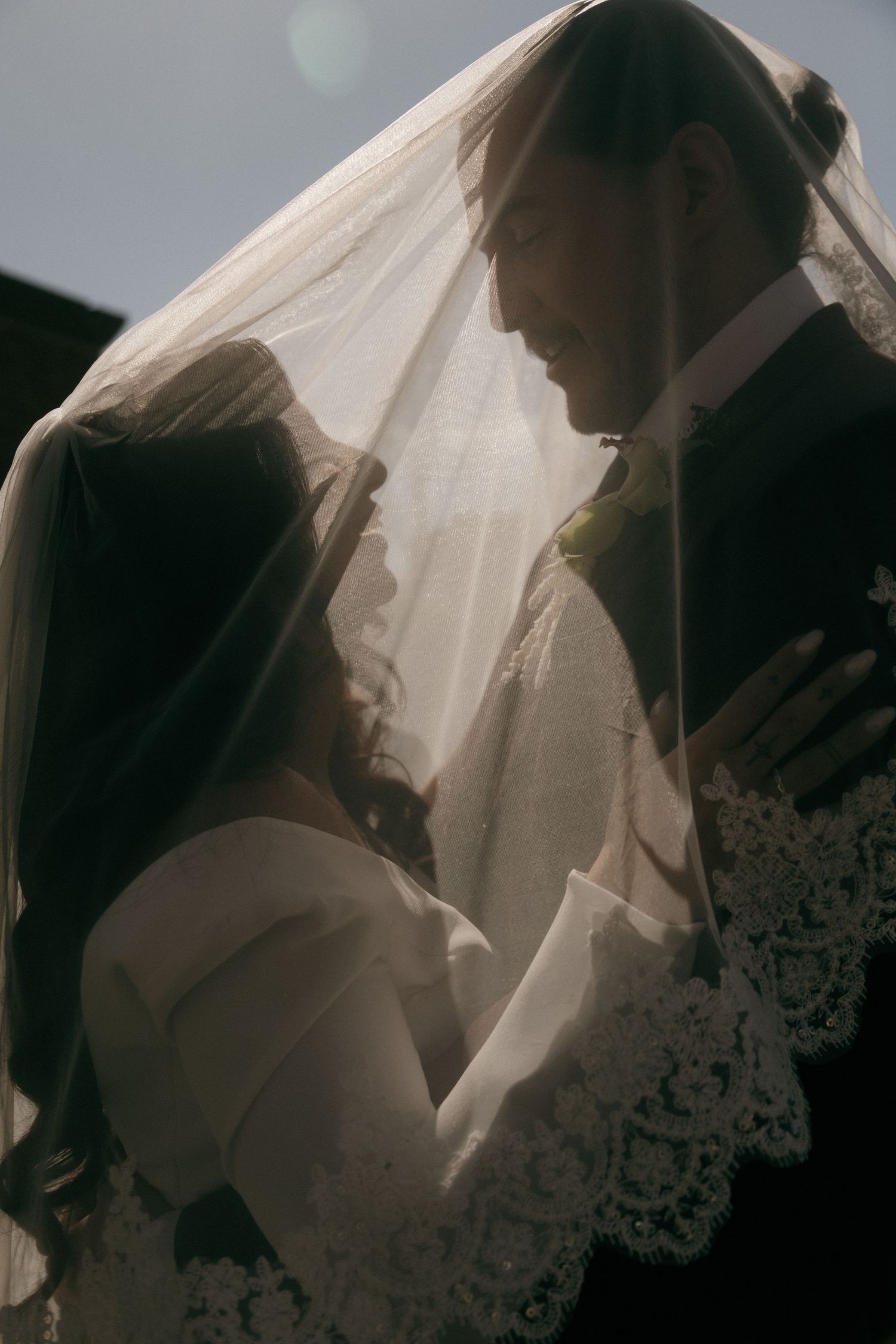 Romantic bride and groom portrait under lace veil backlit by soft sunlight.