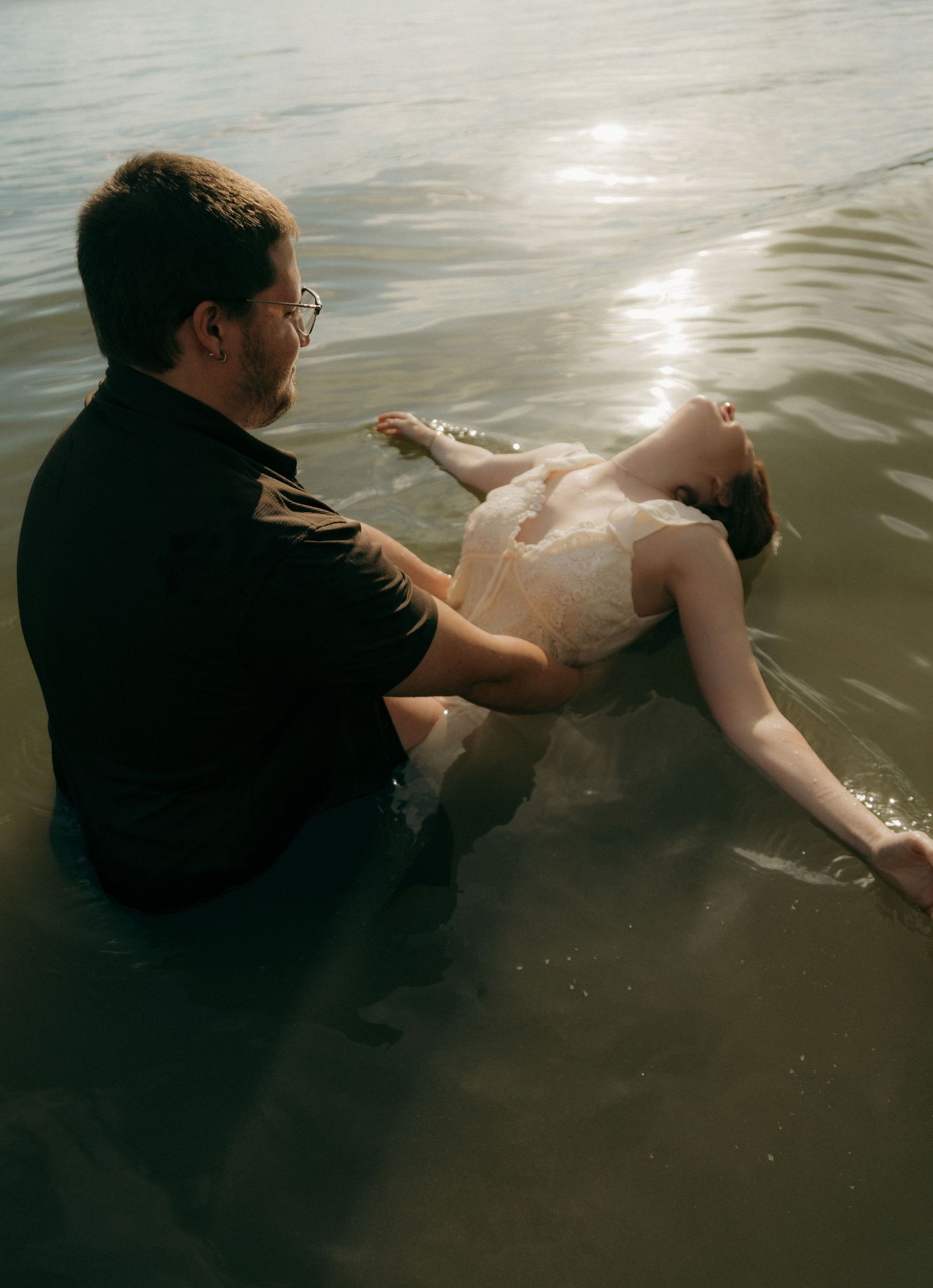 Groom in black shirt holding bride in white dress as she floats in shallow sunlit water.