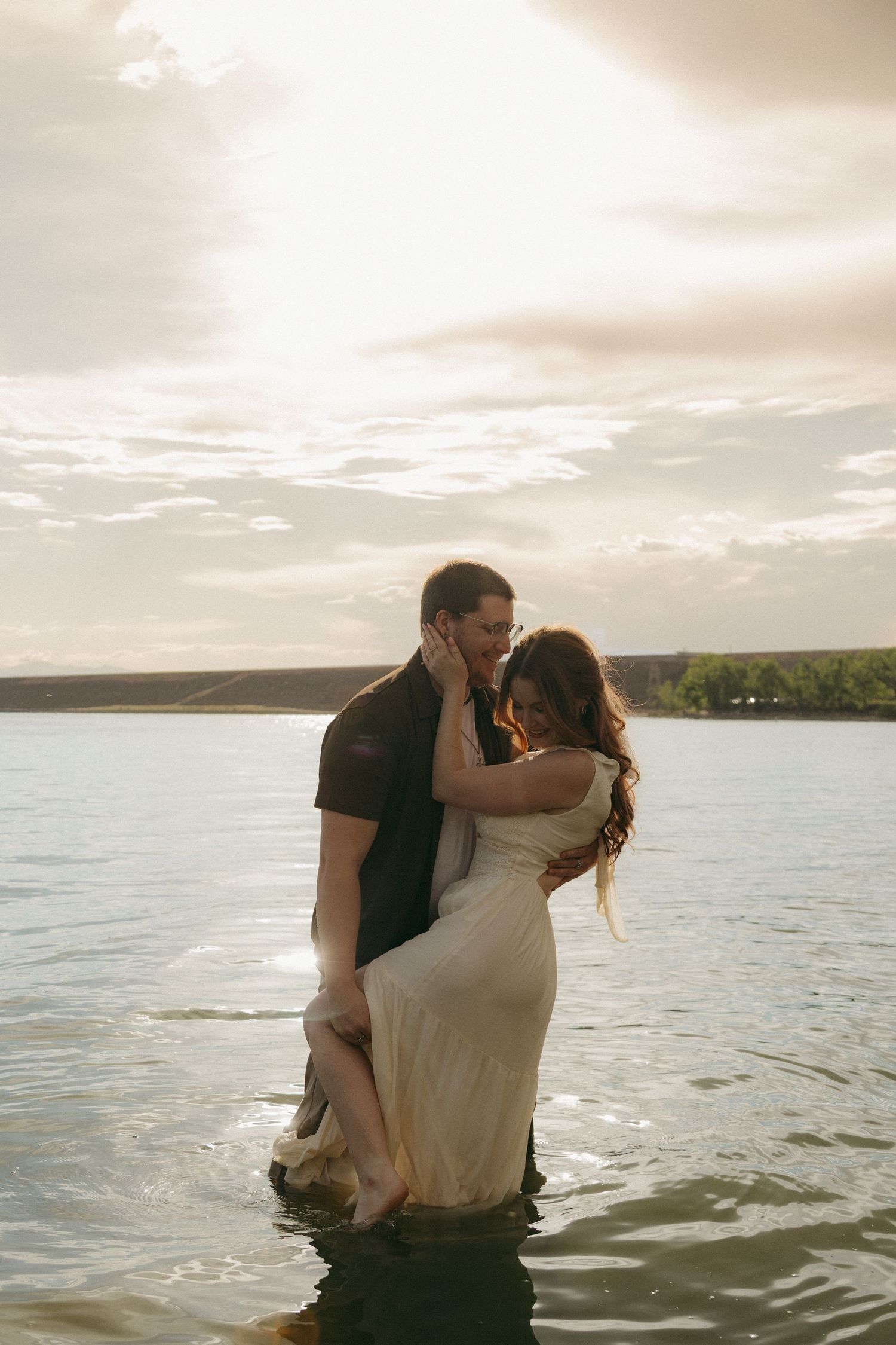Romantic couple embracing in a lake at sunset, woman in white dress and man in black shirt.