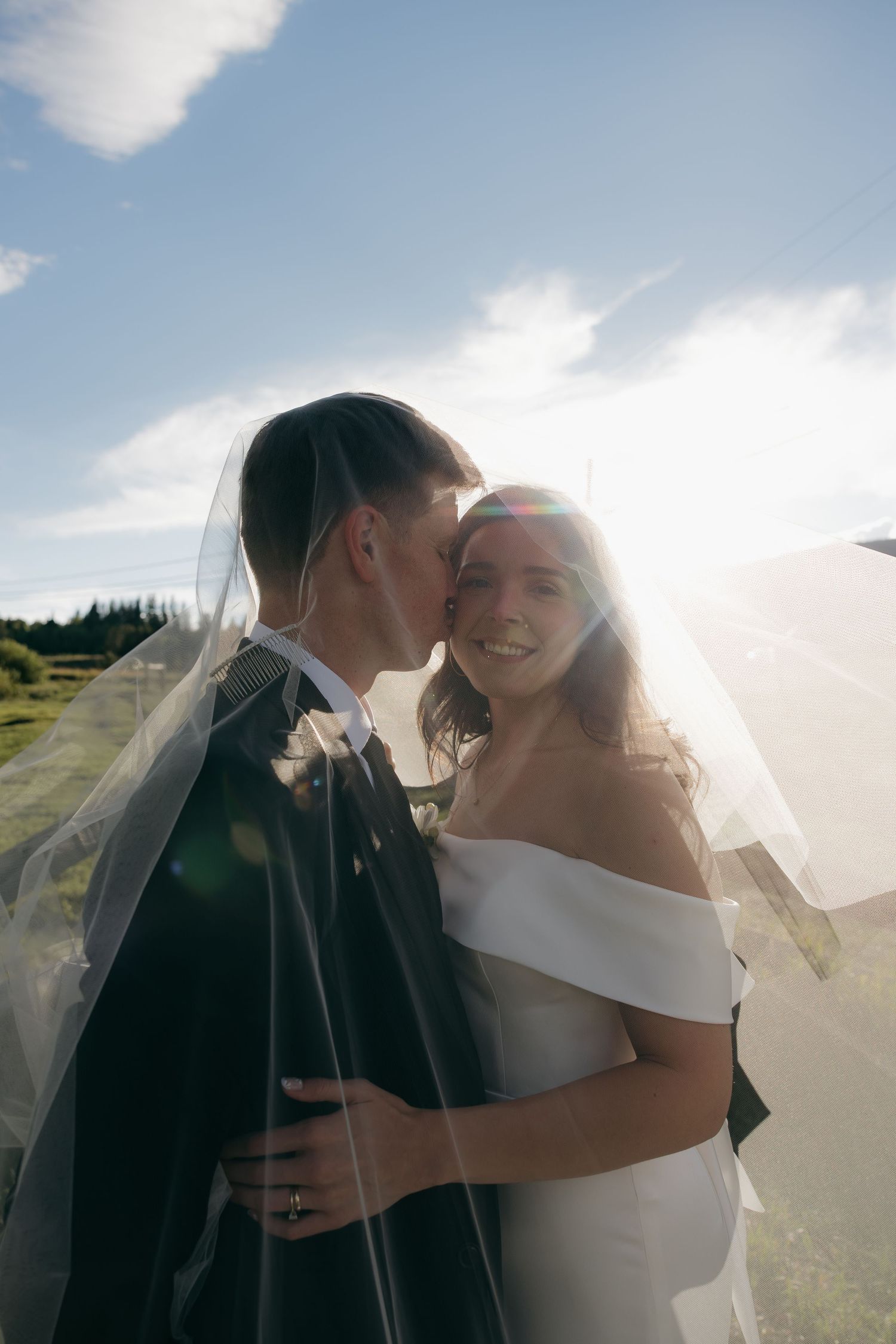 Groom kissing smiling bride on cheek under sheer veil with bright blue sky background.