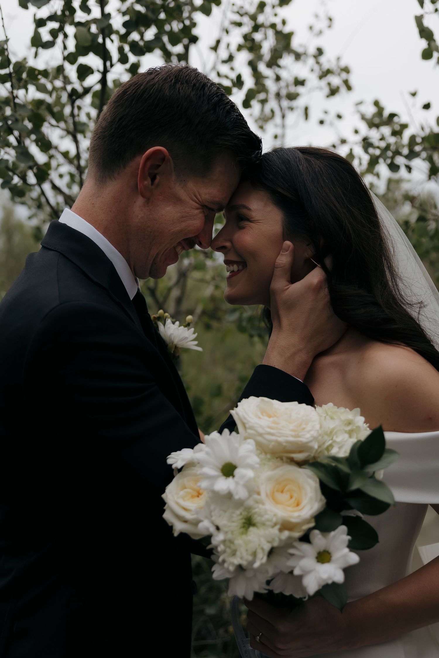 Smiling bride and groom touch foreheads tenderly while bride holds white floral bouquet outdoors.
