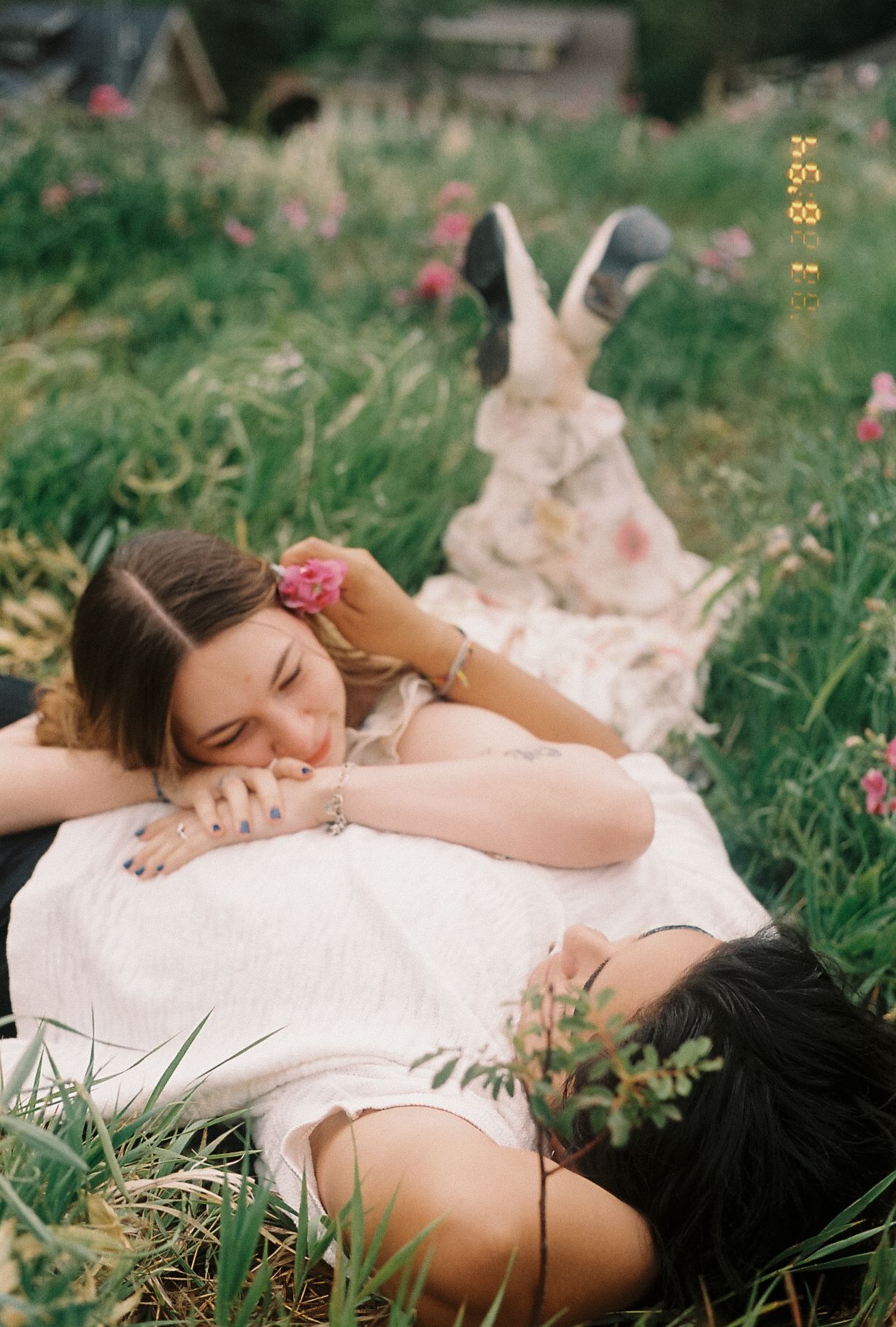 Two people lying in a flower field, one resting head on the other's chest, with a pink flower in their hair.