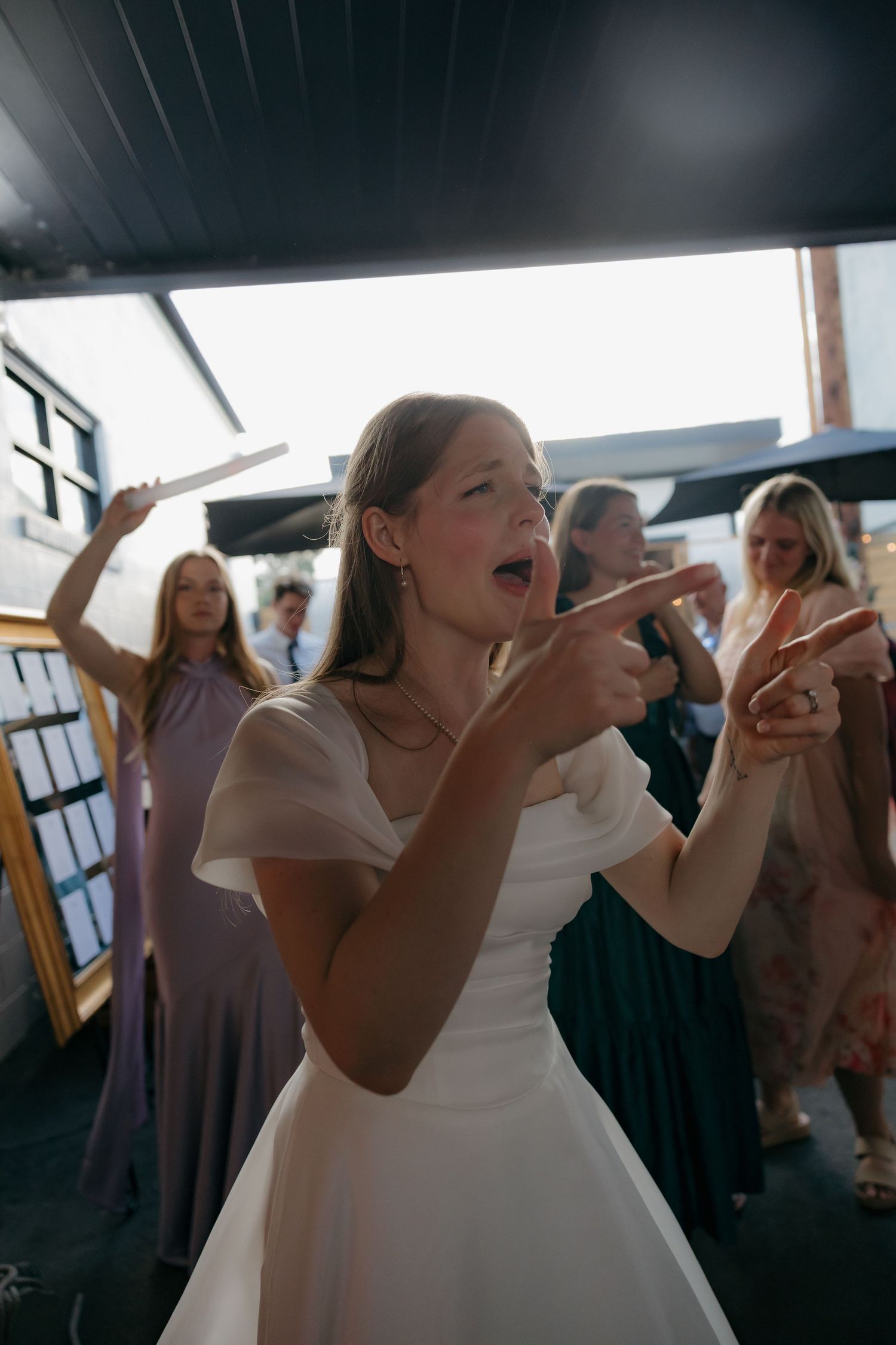 Bride in white dress dancing enthusiastically with wedding guests at outdoor reception celebration.