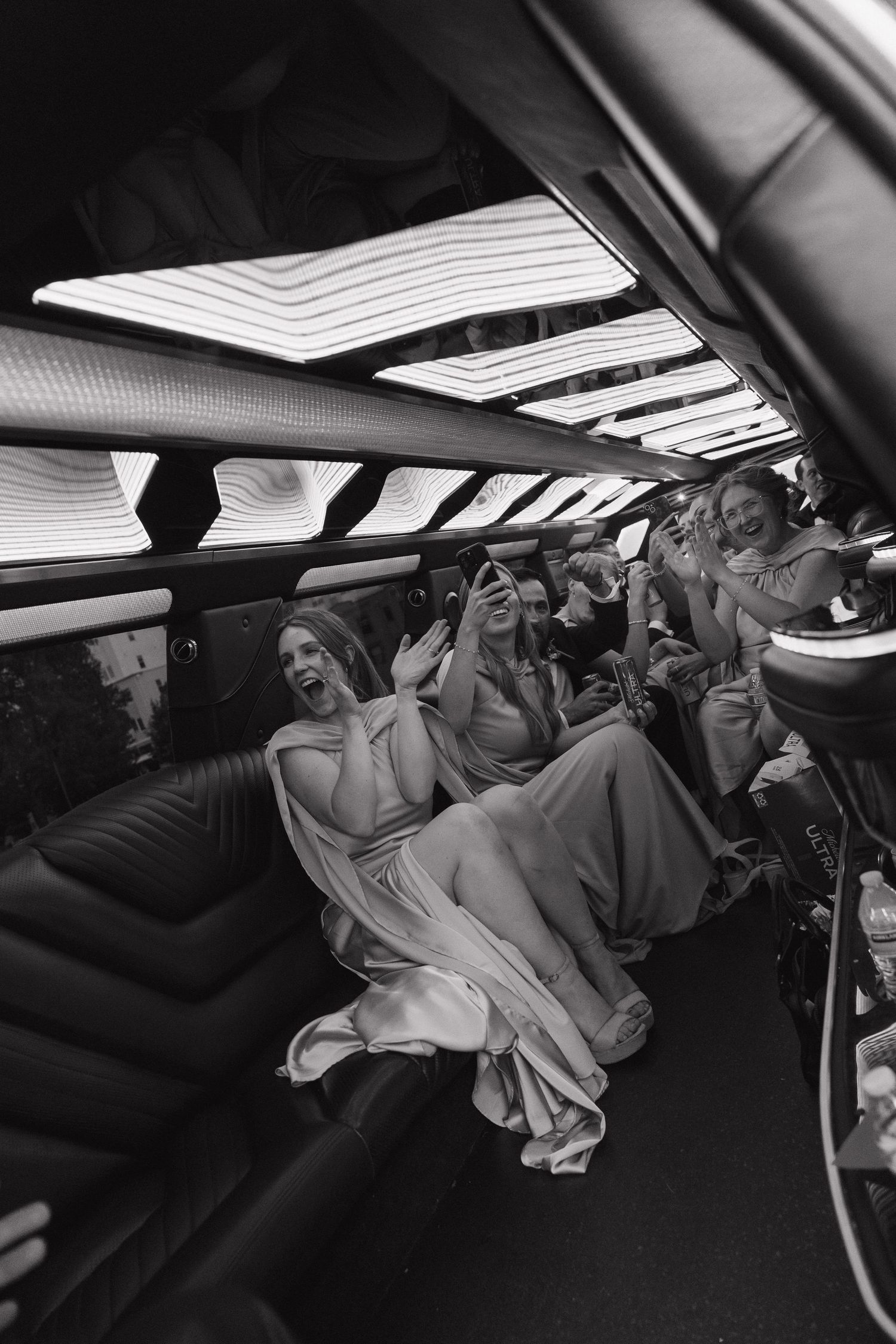 Bridesmaids in formal gowns laugh and take photos inside a limousine in a black and white shot.