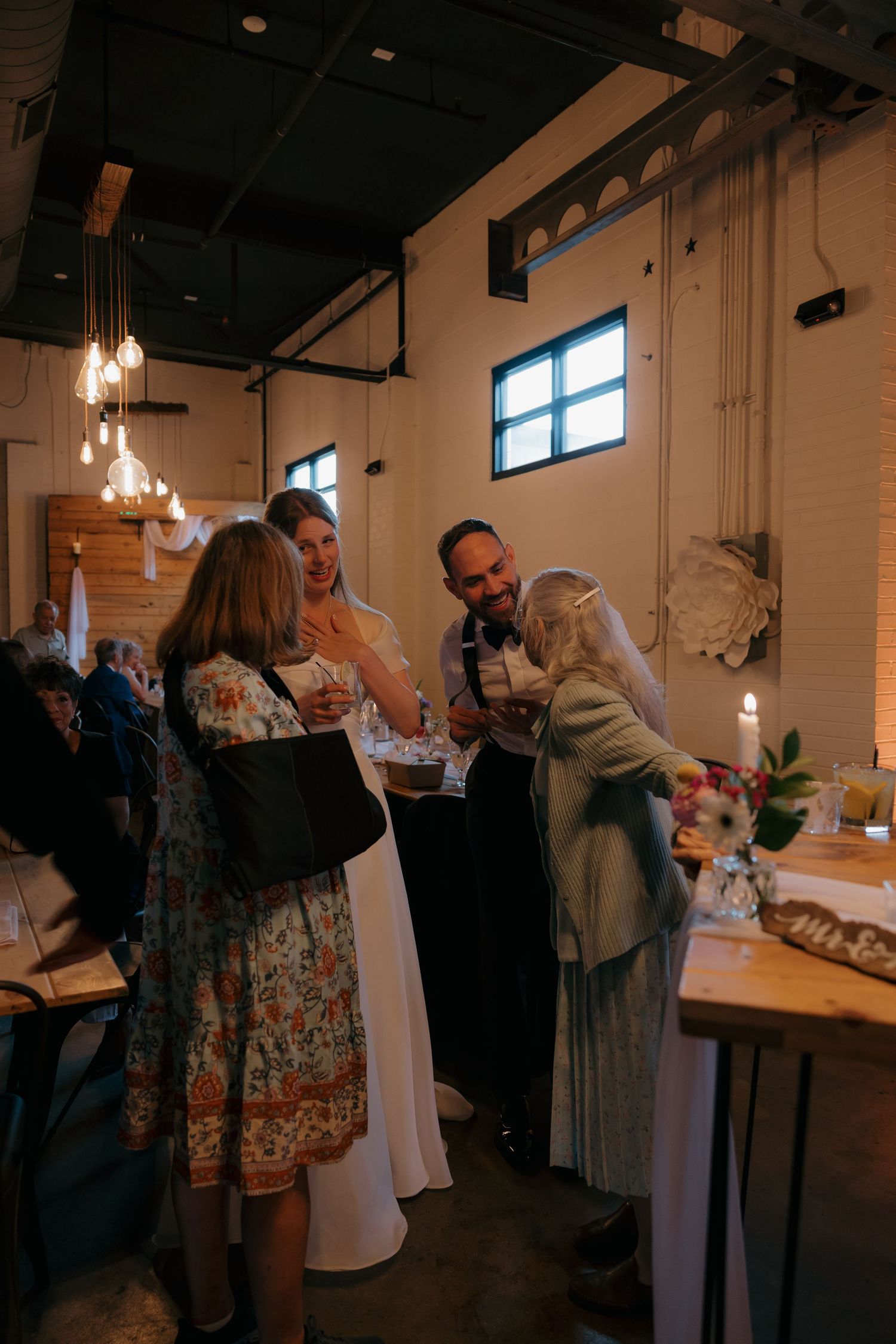 Wedding guests congratulate the bride and groom inside a warmly lit rustic industrial venue.