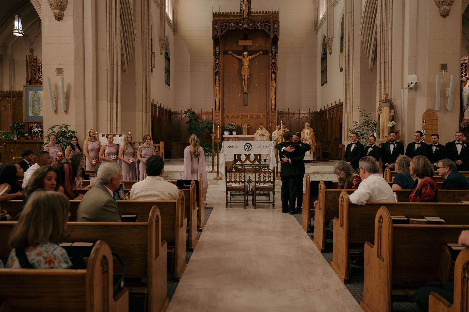 Wedding ceremony in ornate Catholic church with guests seated in wooden pews facing altar.