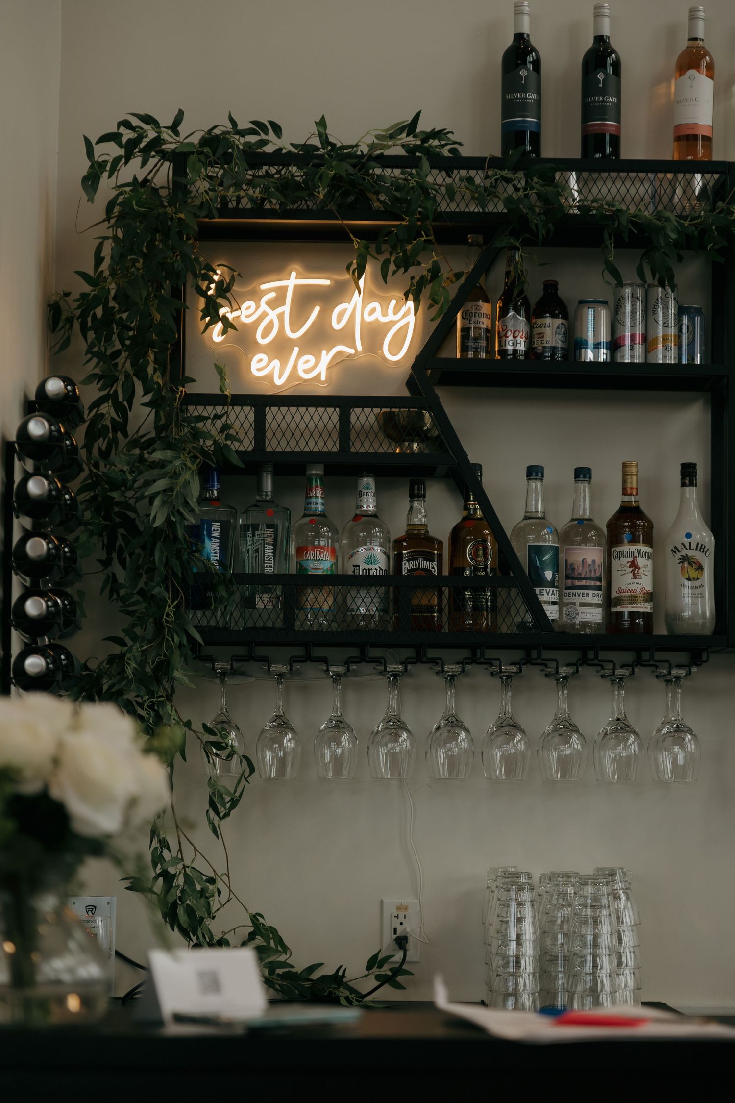 A wedding bar shelf with neon 'best day ever' sign, greenery, wine glasses, and assorted liquor bottles.