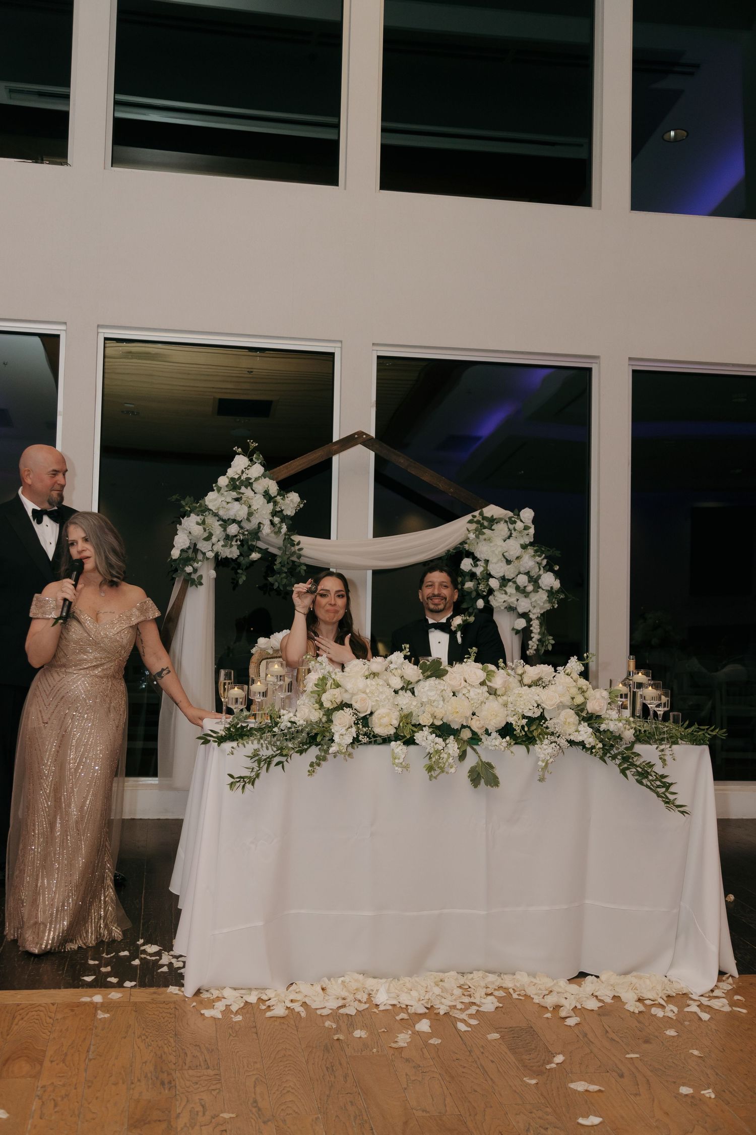 Bride and groom sit at floral sweetheart table with white roses while guests give wedding toast.
