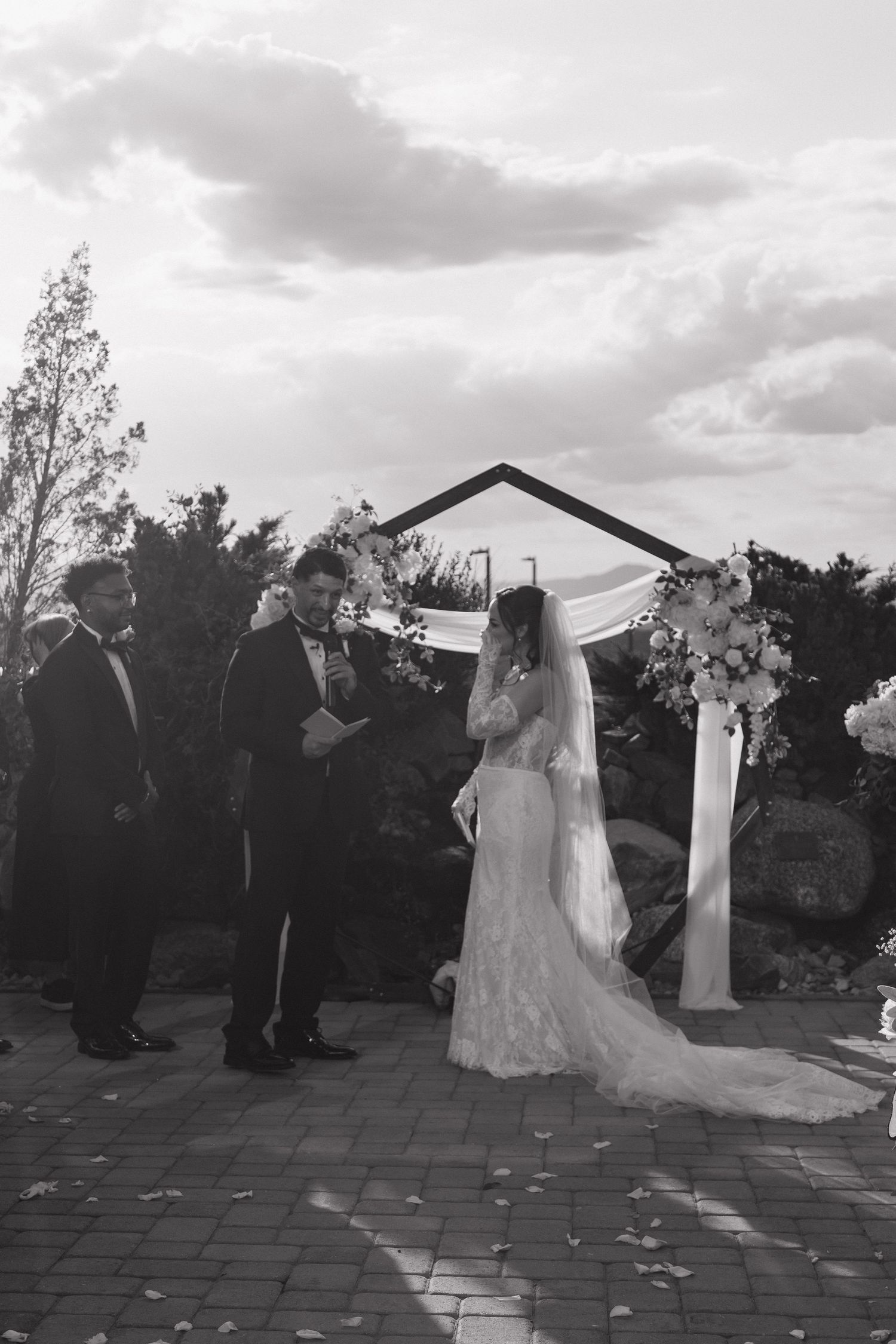 Bride and groom exchange vows at outdoor wedding ceremony beneath floral arch in black and white photo.