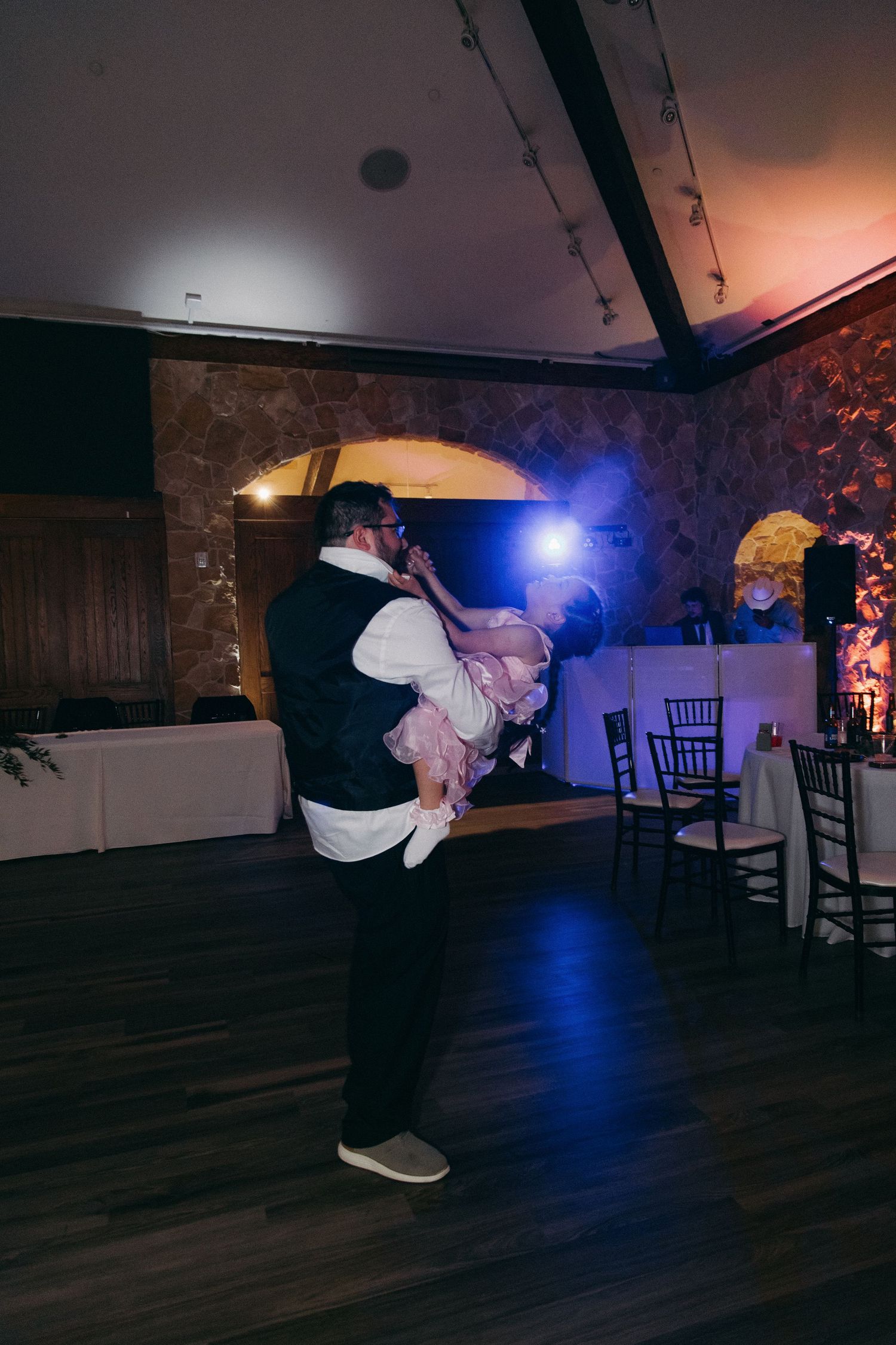 Groom lifting a young flower girl in a pink dress on the dimly lit wedding reception dance floor.