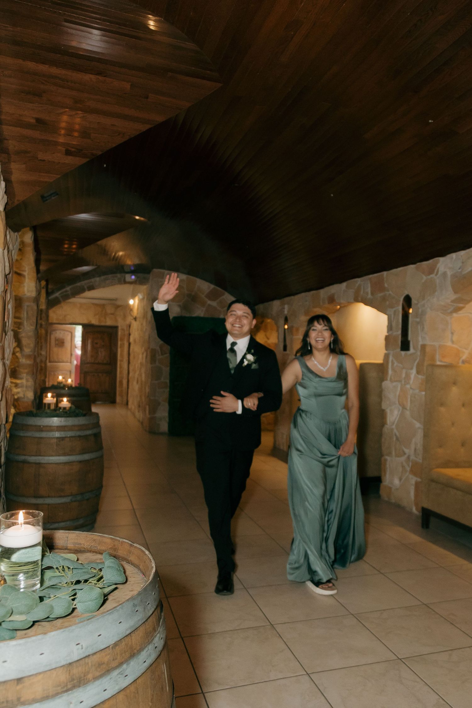 Groom in tuxedo waves while escorted by woman in sage gown through venue corridor.