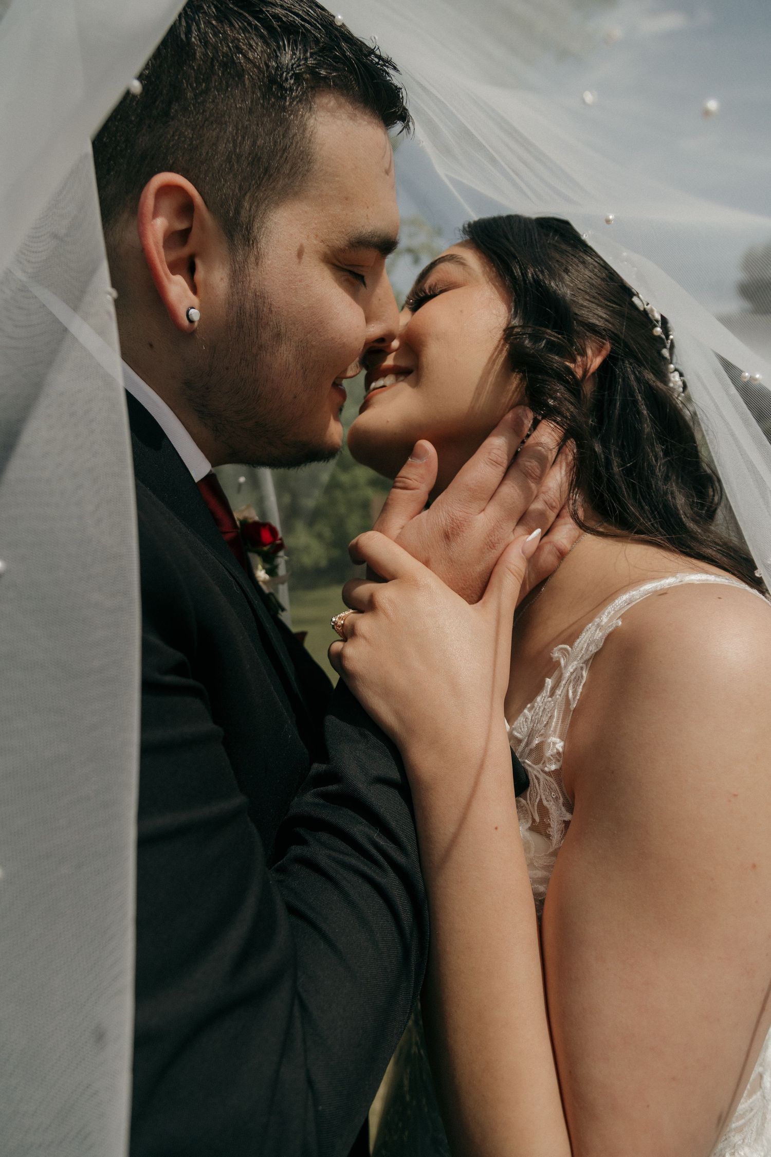 Bride and groom sharing a romantic almost-kiss under a flowing veil in outdoor wedding portrait.