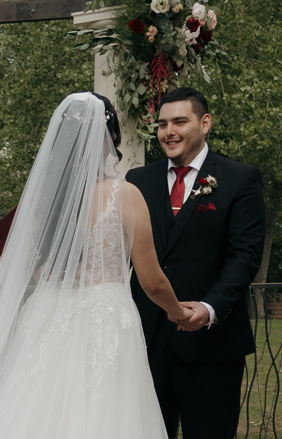 Smiling groom in black suit with red tie holds hands with bride in white lace gown and veil at outdoor wedding ceremony.