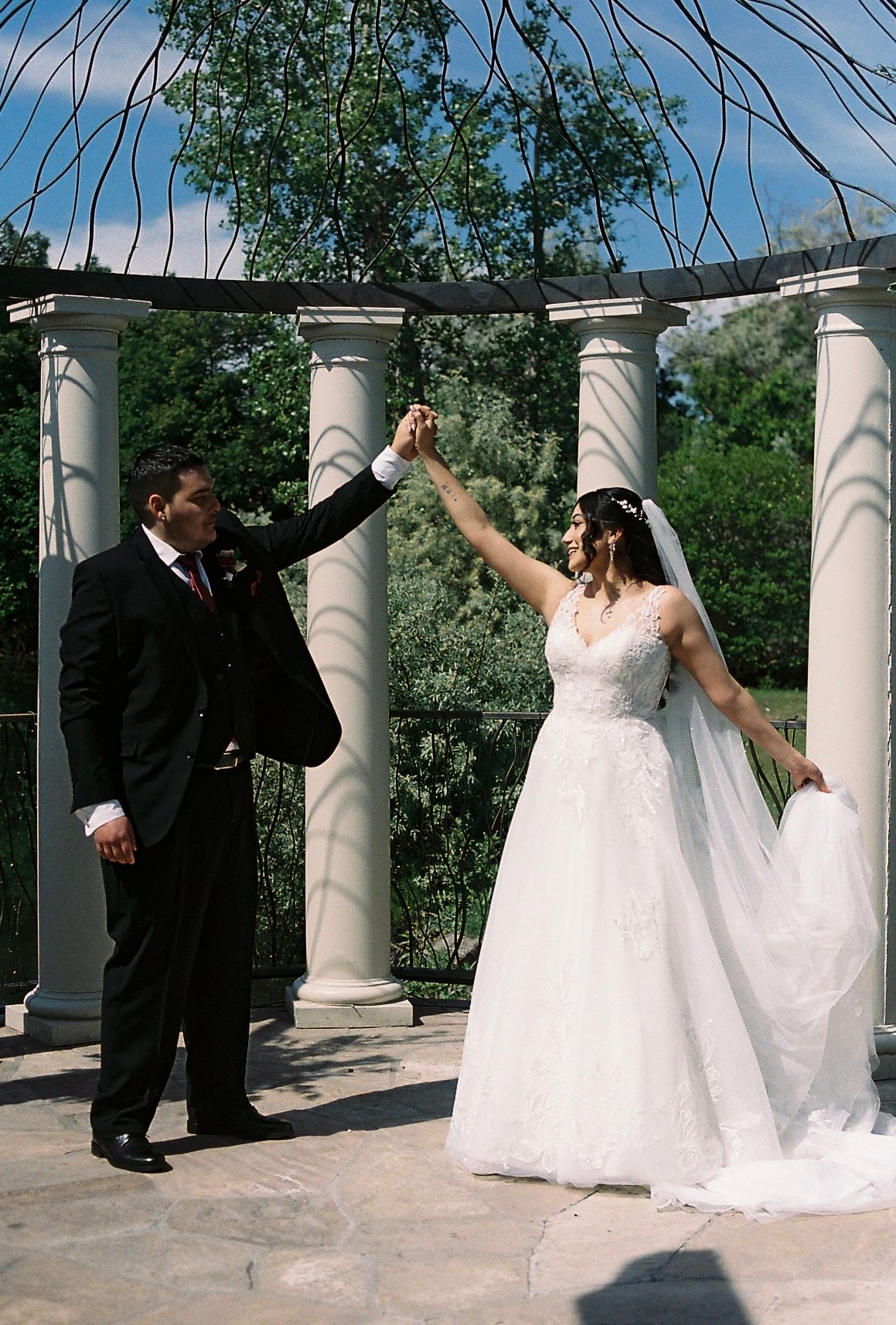 Bride and groom dancing joyfully under white pergola columns during outdoor wedding celebration.