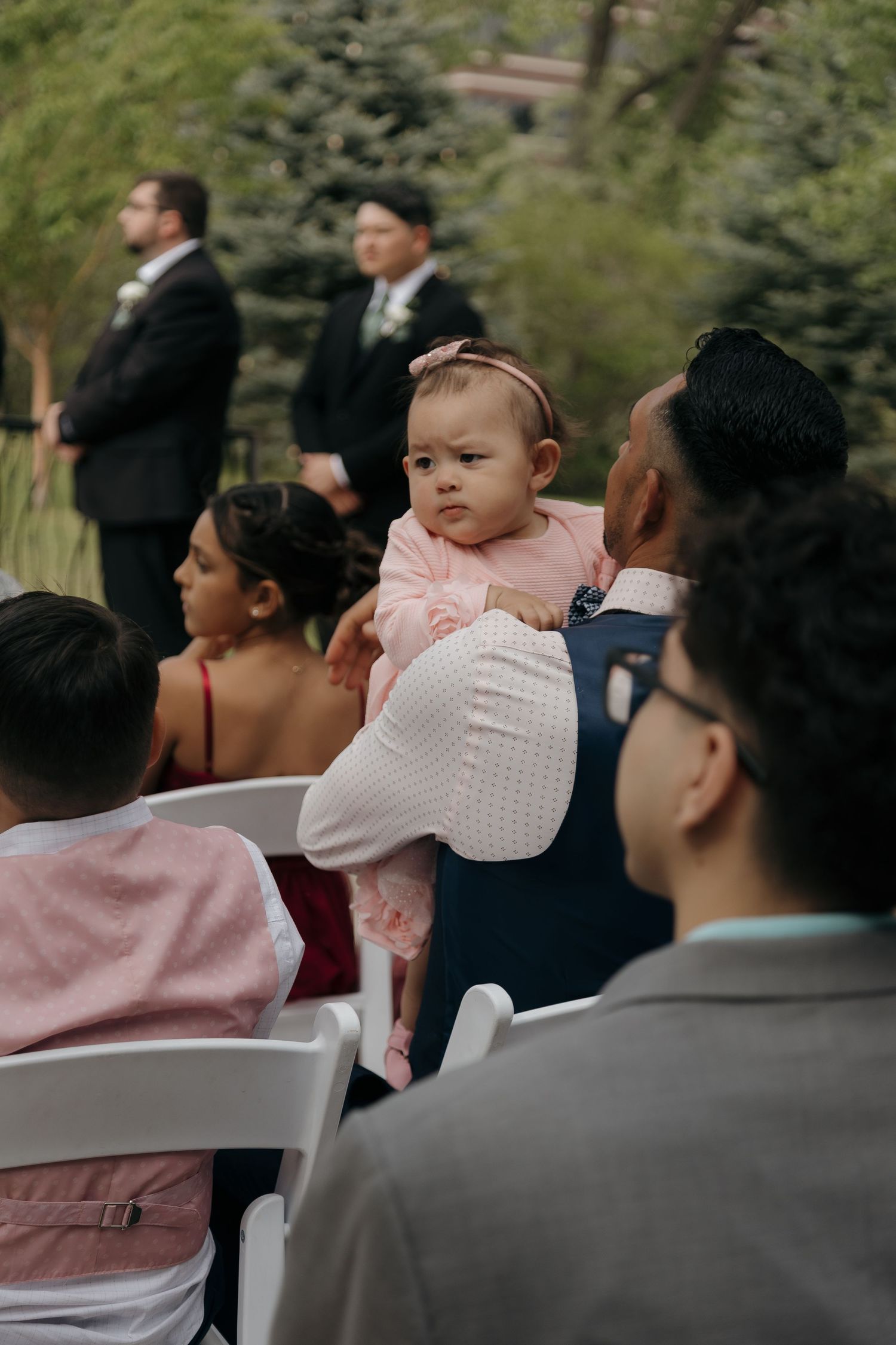 Adorable baby in pink outfit held by guest while watching wedding ceremony from seated audience outdoors.