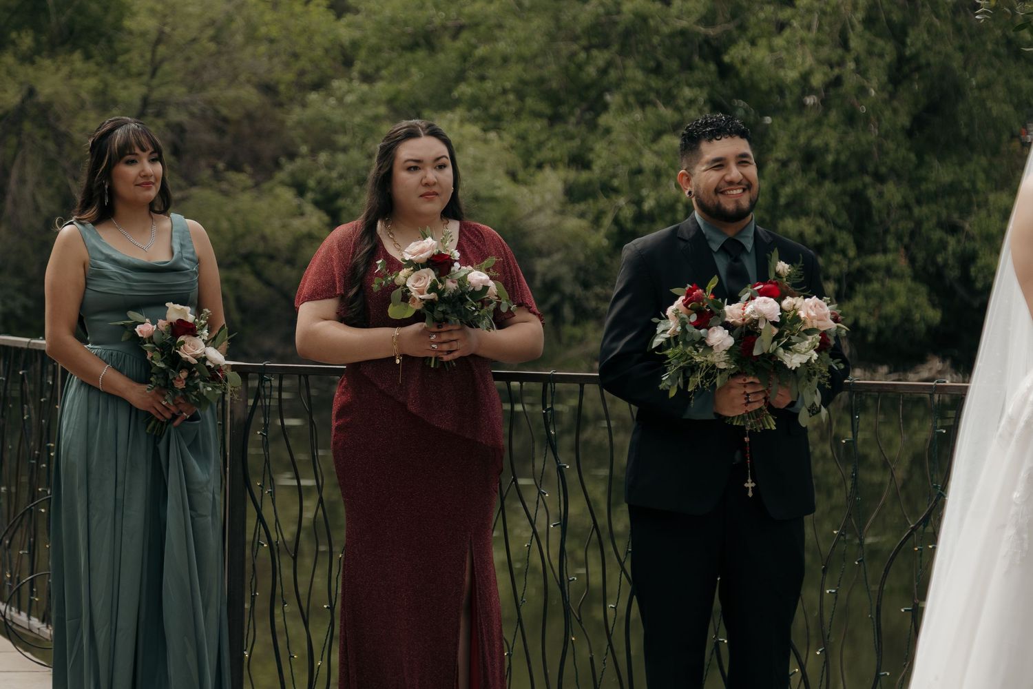 Two bridesmaids in green and burgundy dresses hold bouquets beside groomsman on bridge at wedding.