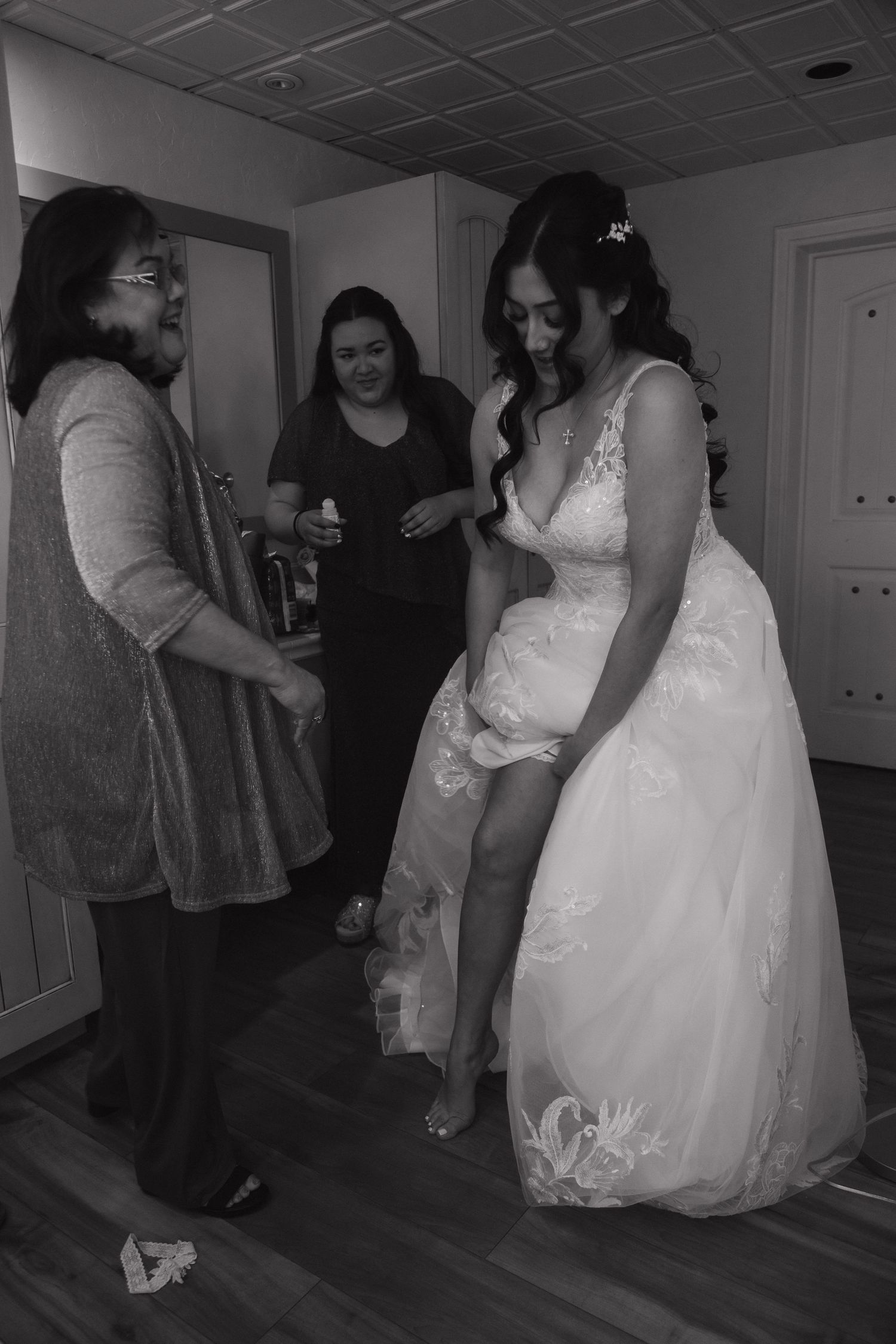 Black and white photo of bride in lace wedding gown being helped by family members in a bridal suite.