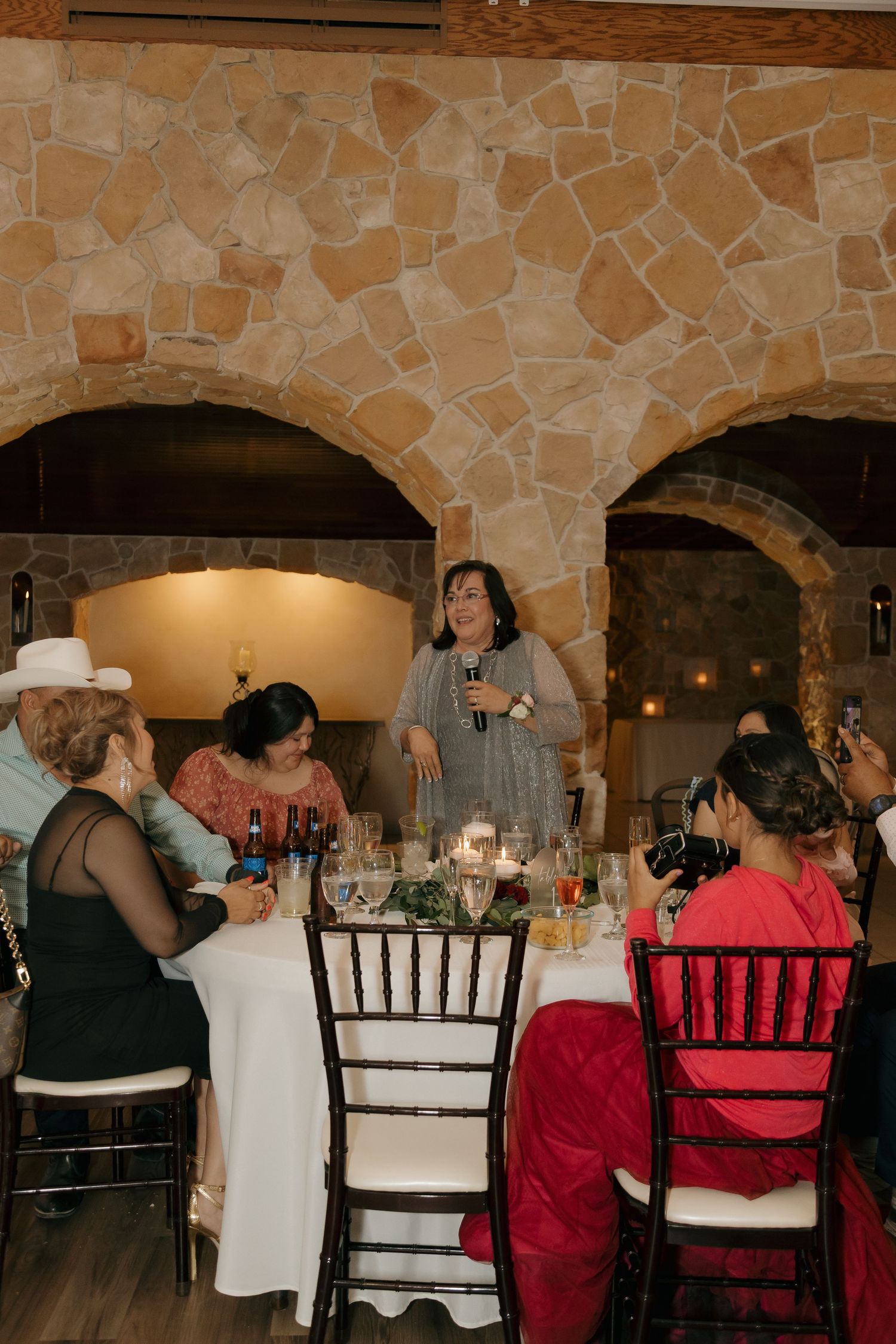 Woman giving a speech at an elegant wedding reception dinner in a rustic stone venue with arched walls.