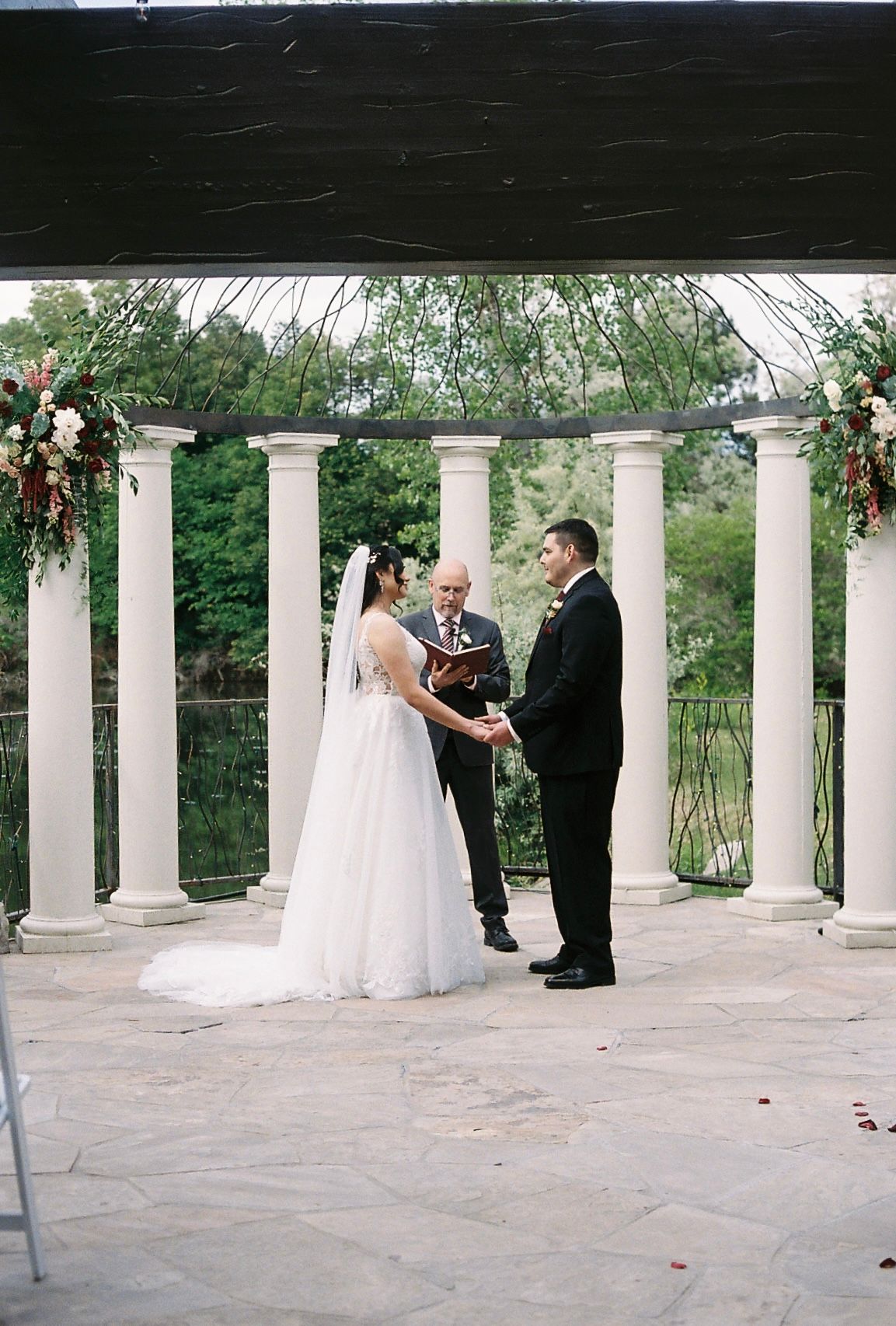 Bride and groom exchange vows at outdoor ceremony beneath white columned pergola adorned with floral arrangements.