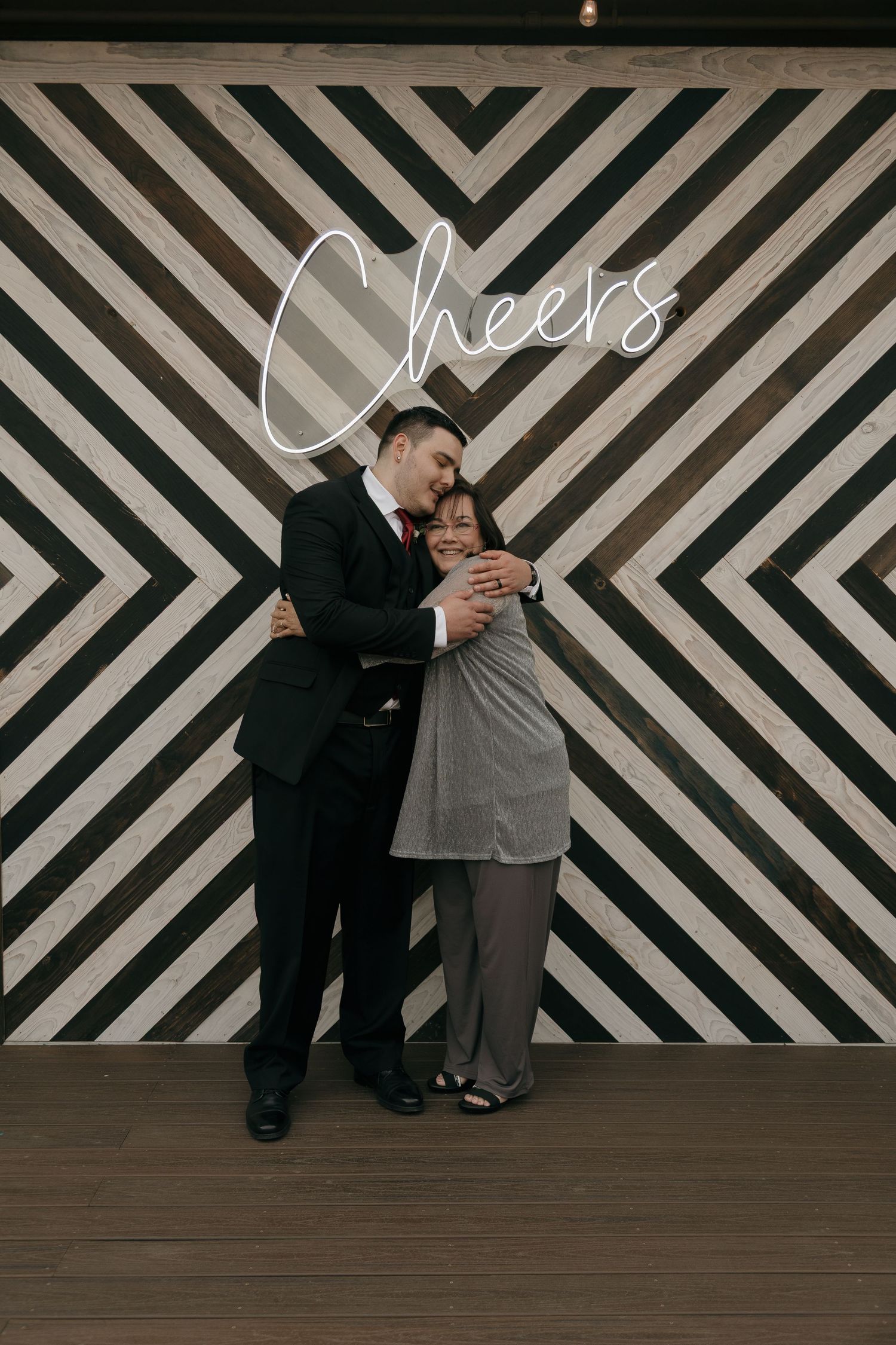 Couple embraces in front of a black and white chevron wall with a glowing Cheers neon sign.