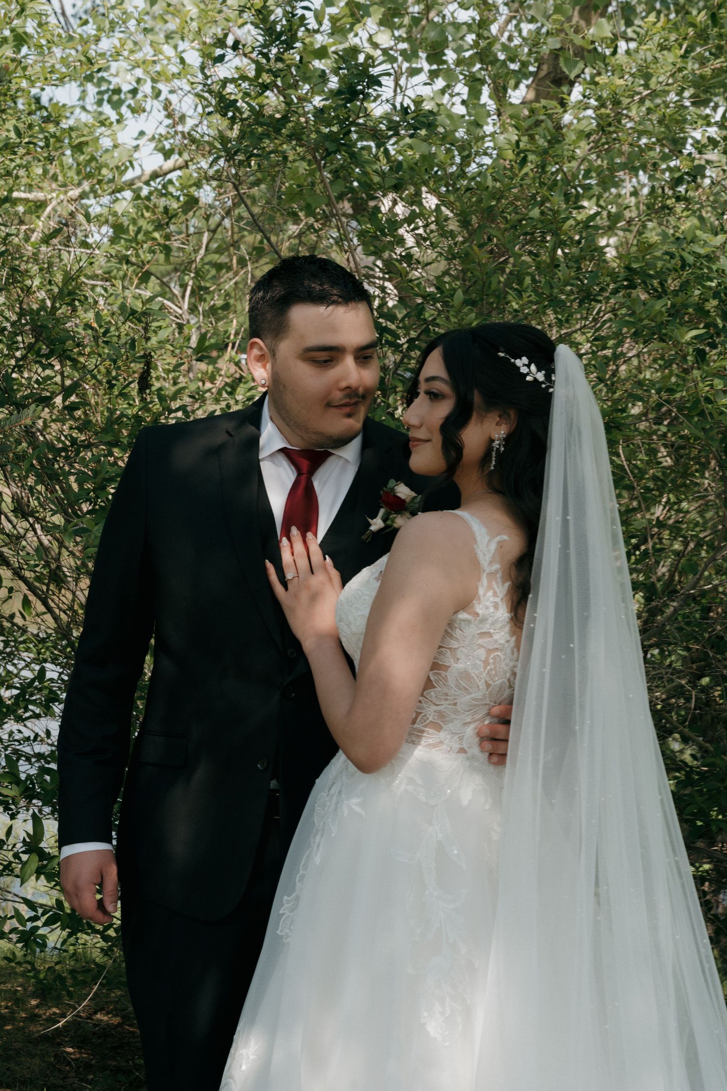 Bride in lace wedding gown and groom in black suit with red tie embracing among green trees.