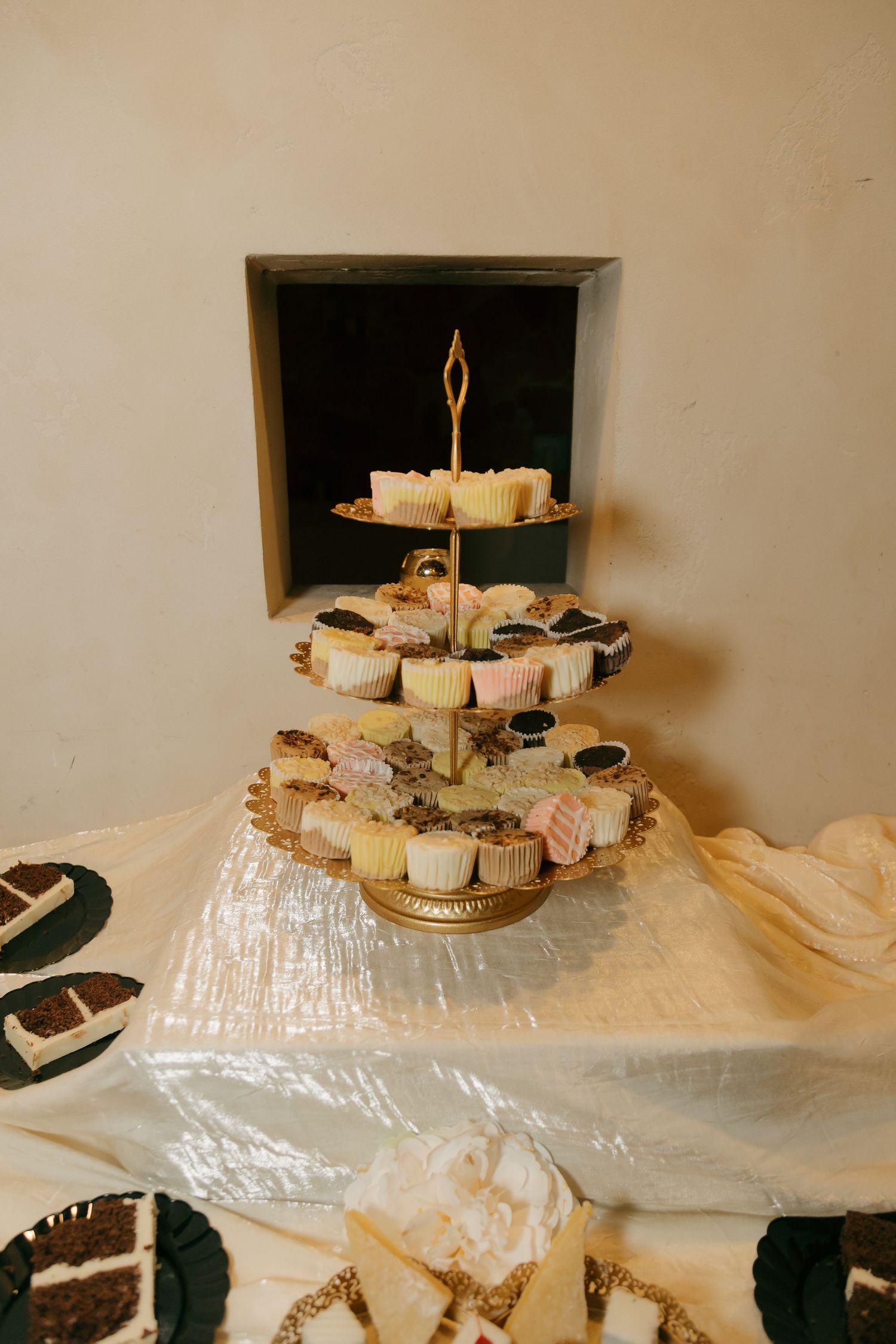Elegant tiered gold cupcake stand displaying assorted wedding desserts on a white draped table.