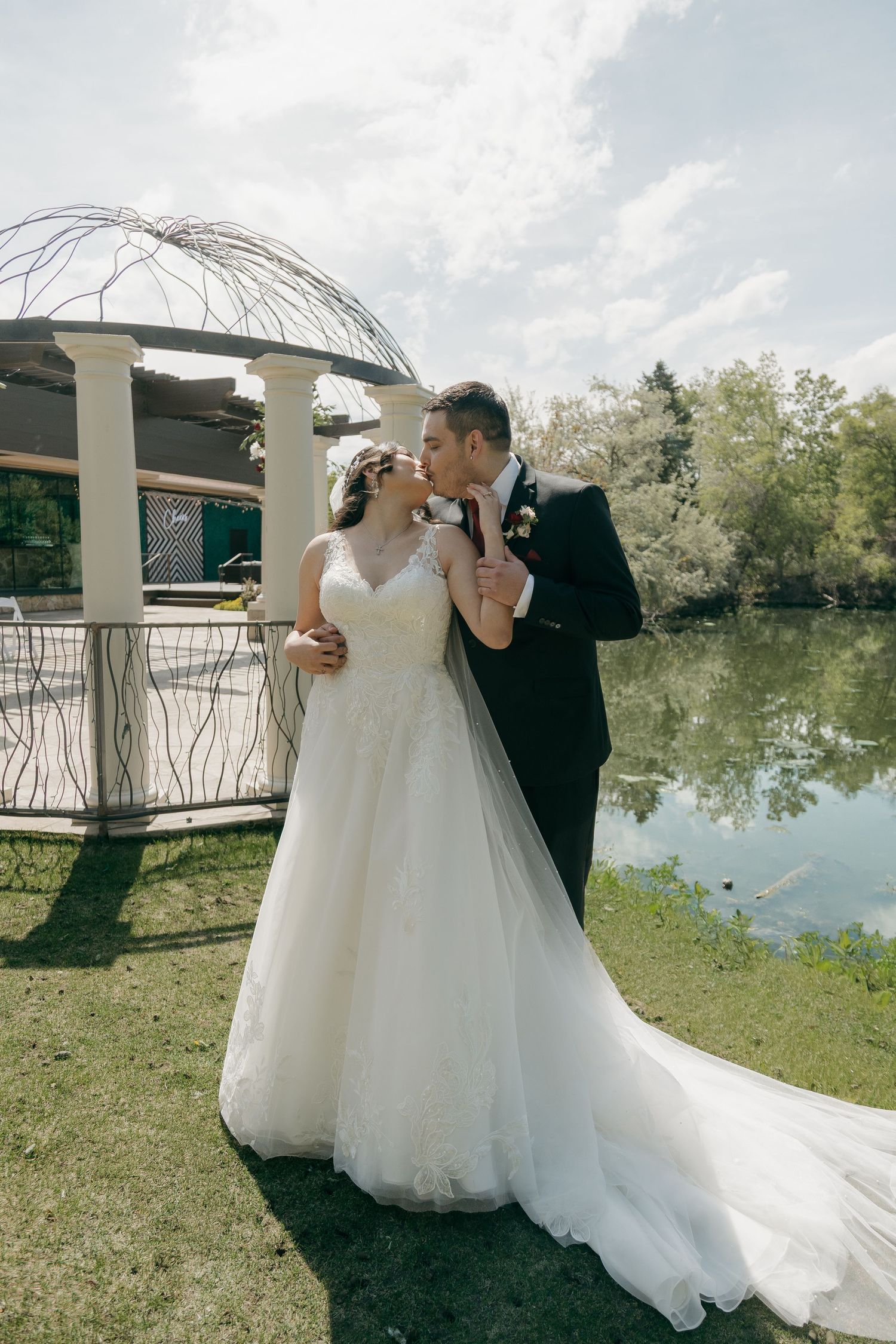 Bride and groom share a romantic kiss near a gazebo by a serene pond on their wedding day.