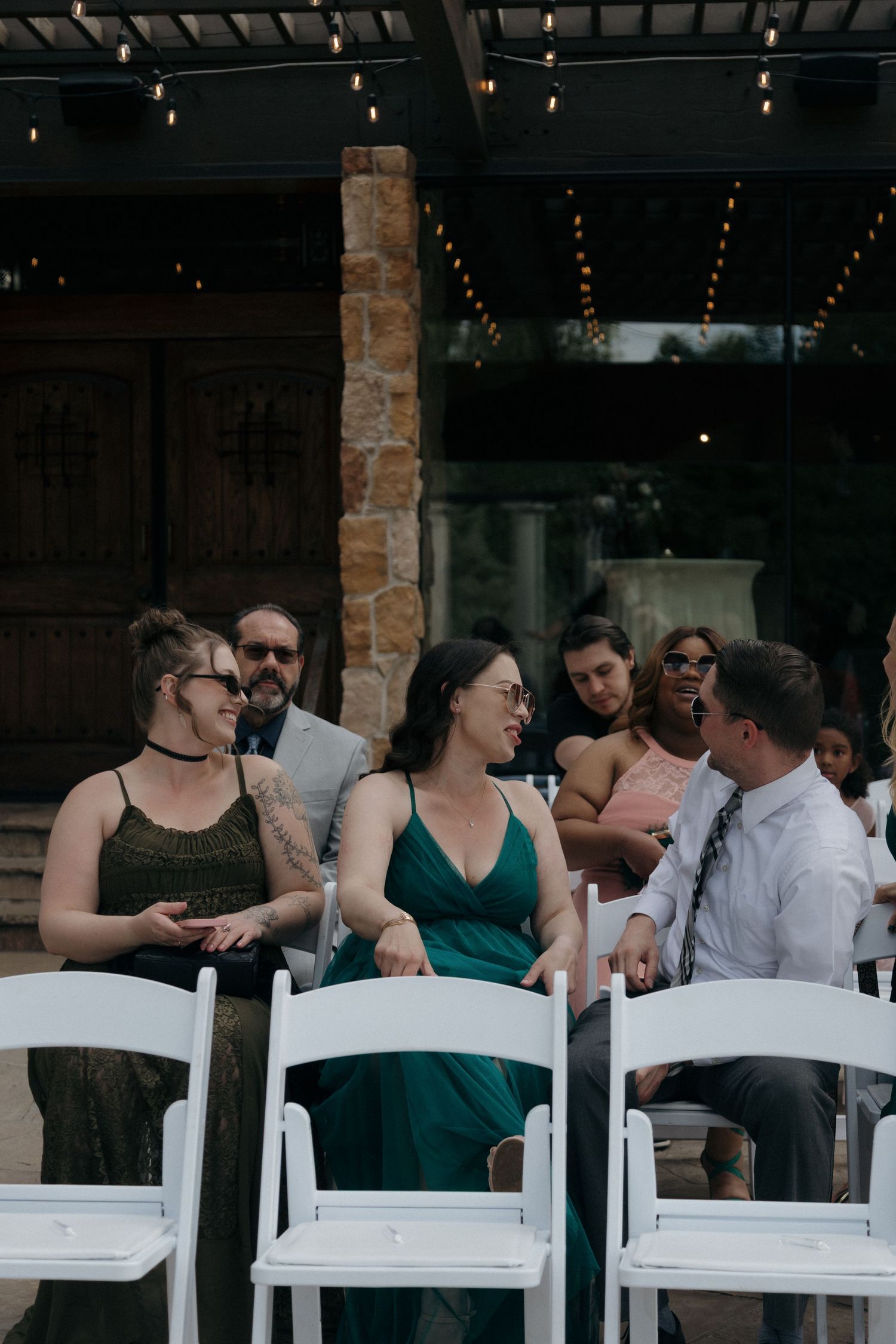 Wedding guests in formal attire chat and laugh while seated in white chairs at an outdoor ceremony.