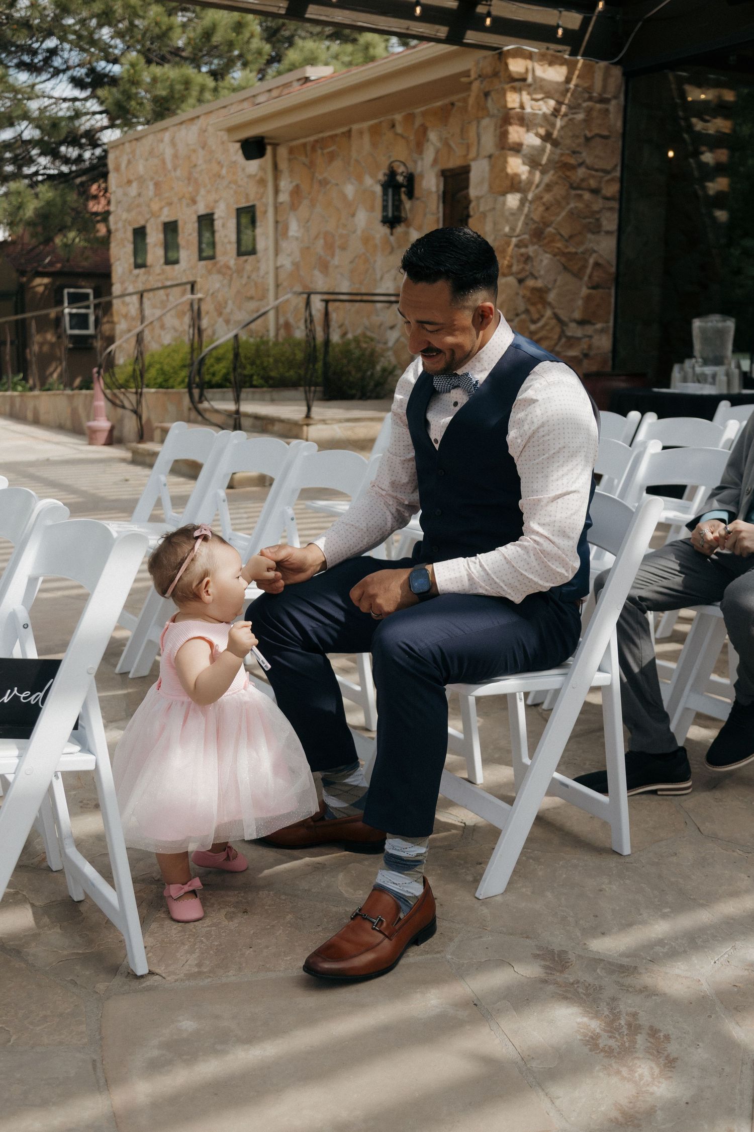 A smiling groom in a navy vest interacts with an adorable toddler girl in a pink dress at an outdoor wedding ceremony.