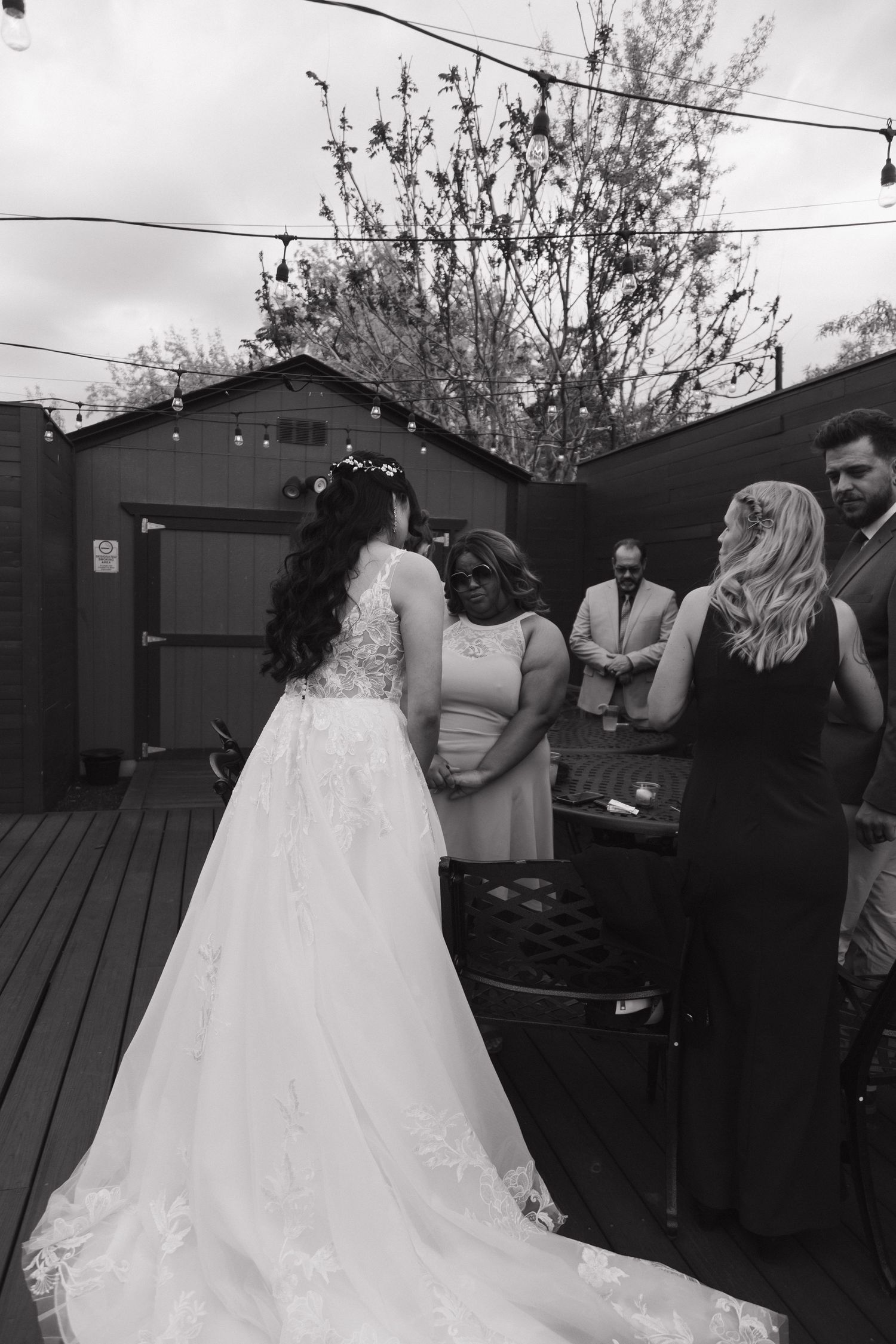 Black and white photo of bride in lace gown speaking with guests during outdoor backyard wedding reception.