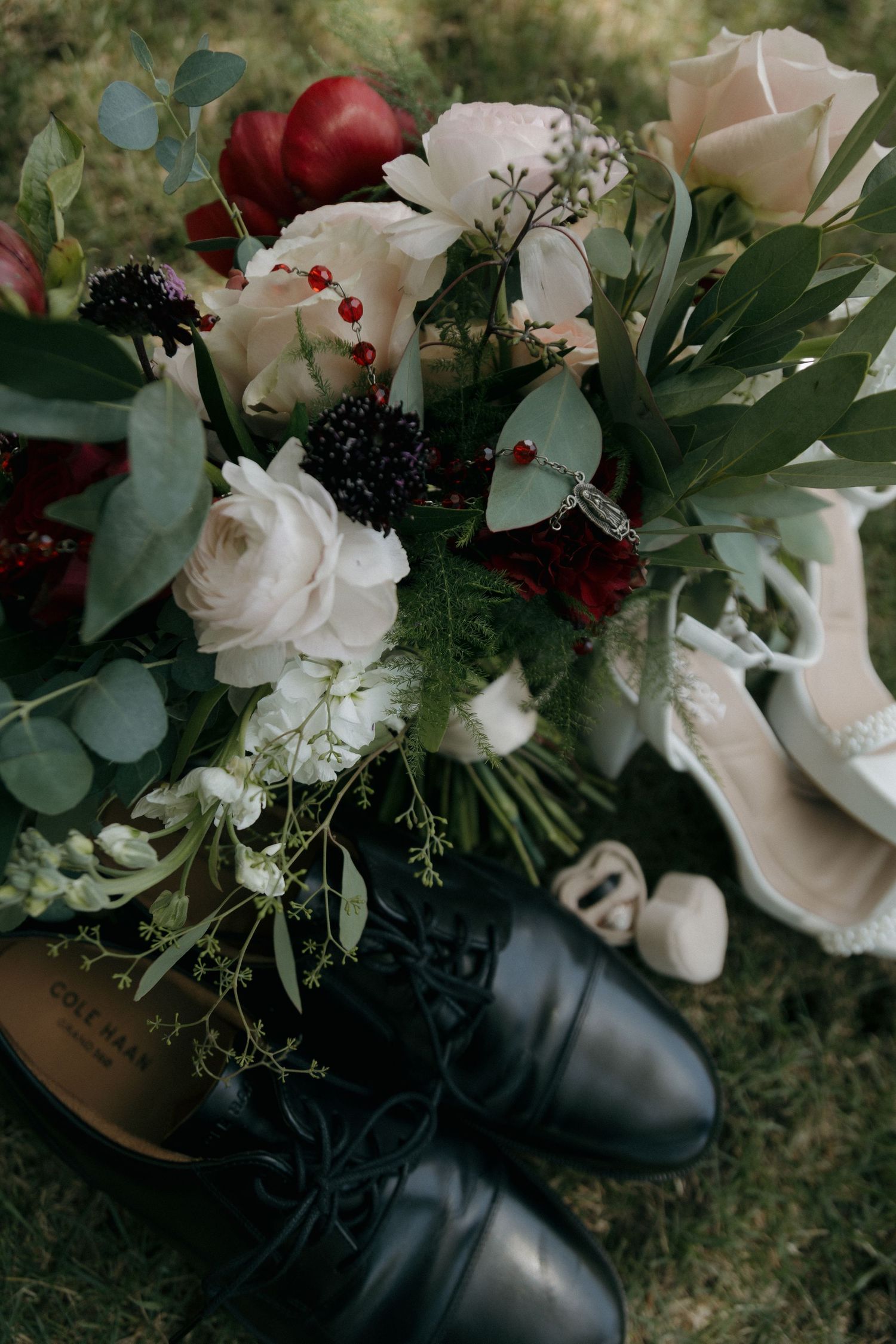 Wedding bouquet with white and red roses beside black groom shoes and white bridal heels on grass.