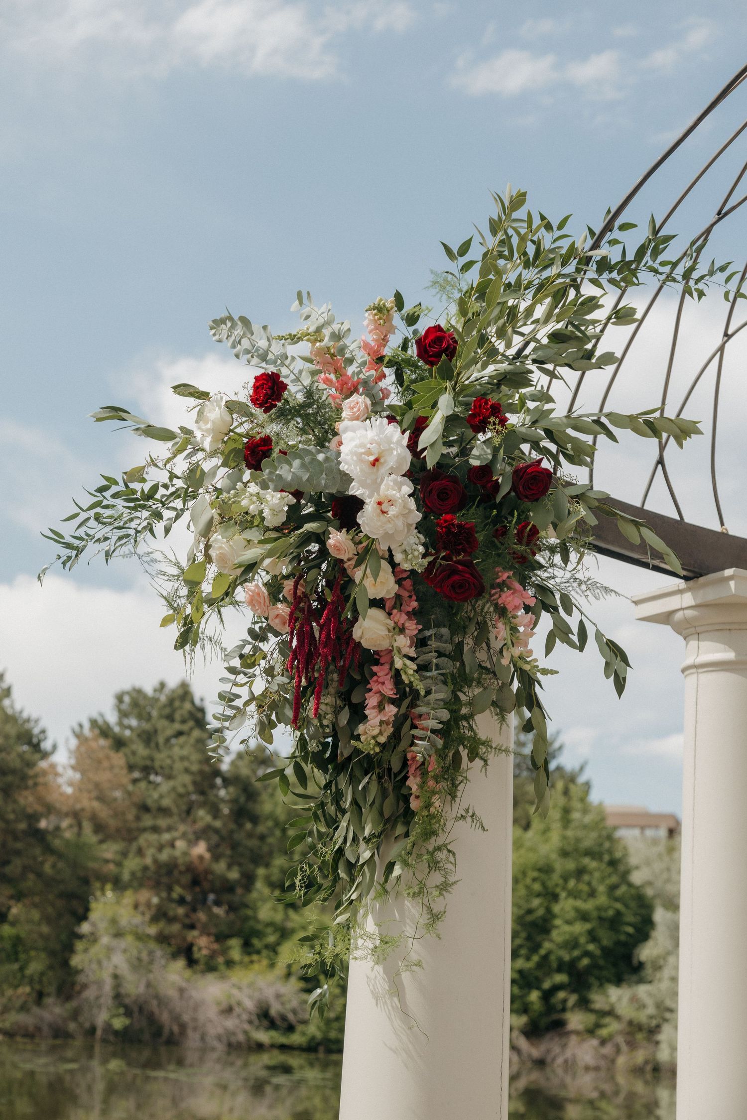 Elegant wedding arch adorned with lush greenery, red roses, blush peonies, and cascading floral arrangements against a blue sky.