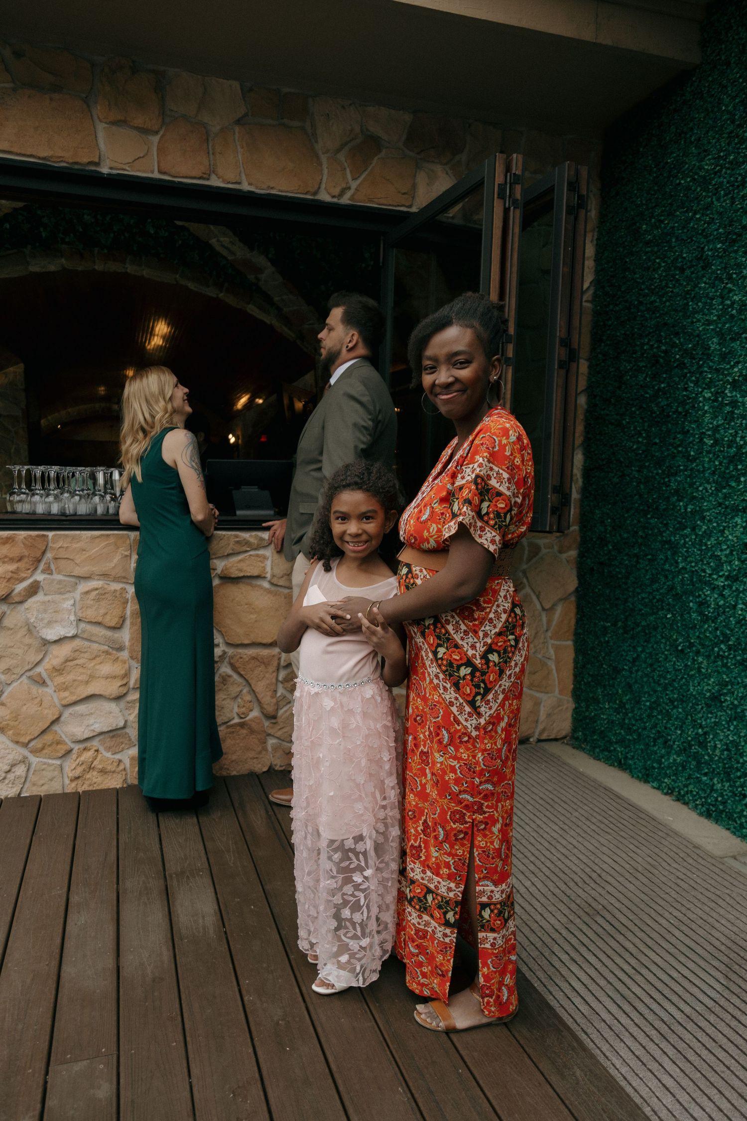 Woman in red floral dress embraces young girl in pink at a wedding reception on a stone venue patio.
