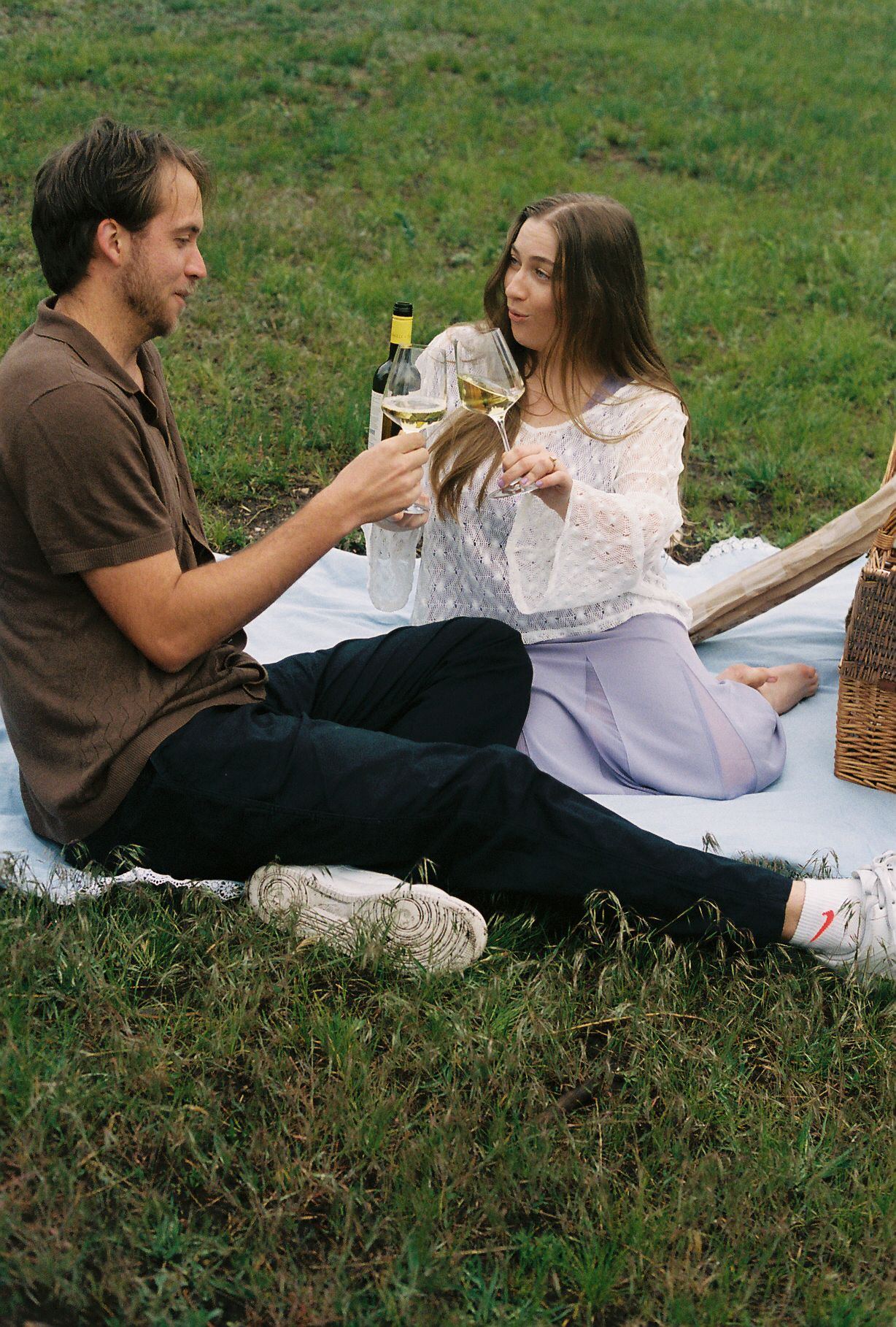 Couple toasting with white wine glasses during a romantic outdoor picnic on a green grass field.
