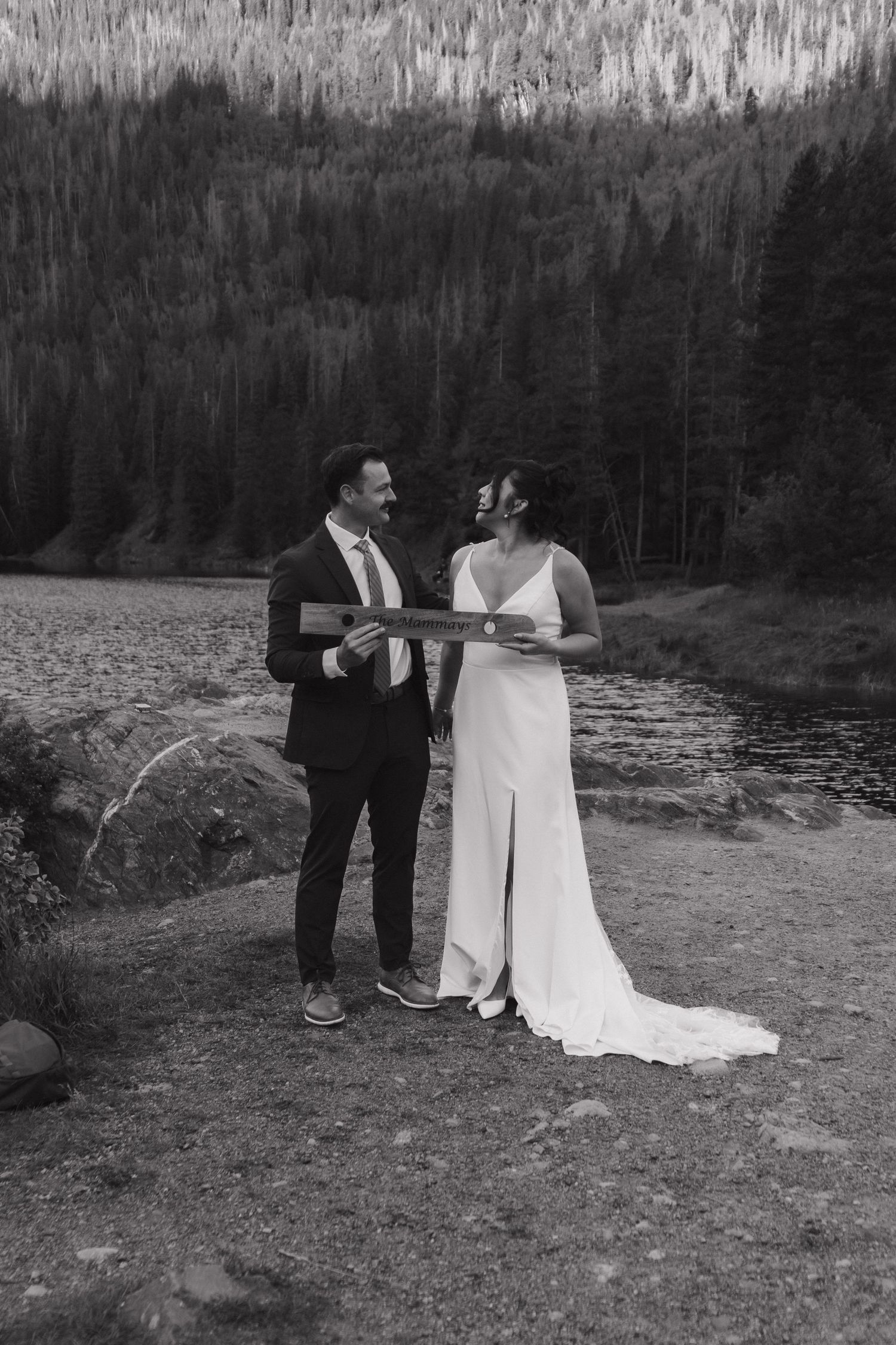 Black and white photo of wedding couple holding wooden sign during lakeside ceremony.