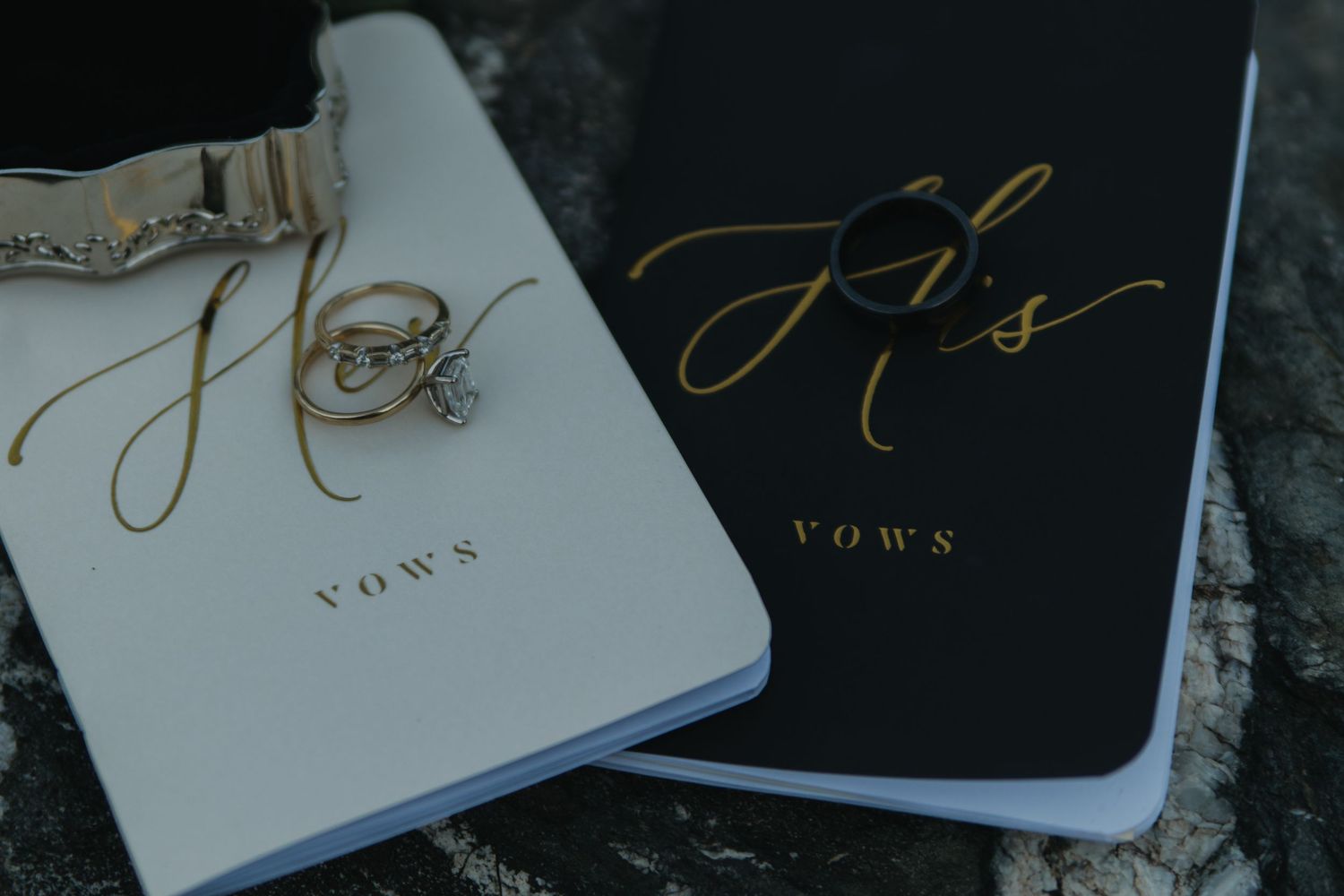 Black and white wedding vow books with gold calligraphy lettering lay on a rustic wooden surface.