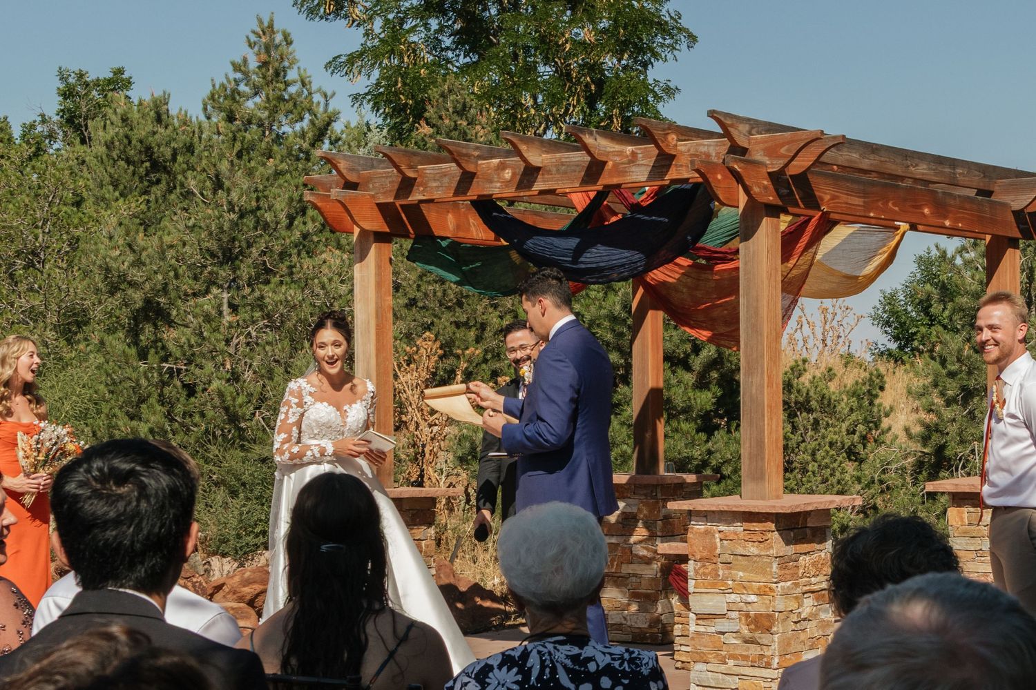 Outdoor wedding ceremony under a wooden pergola with natural landscaping.