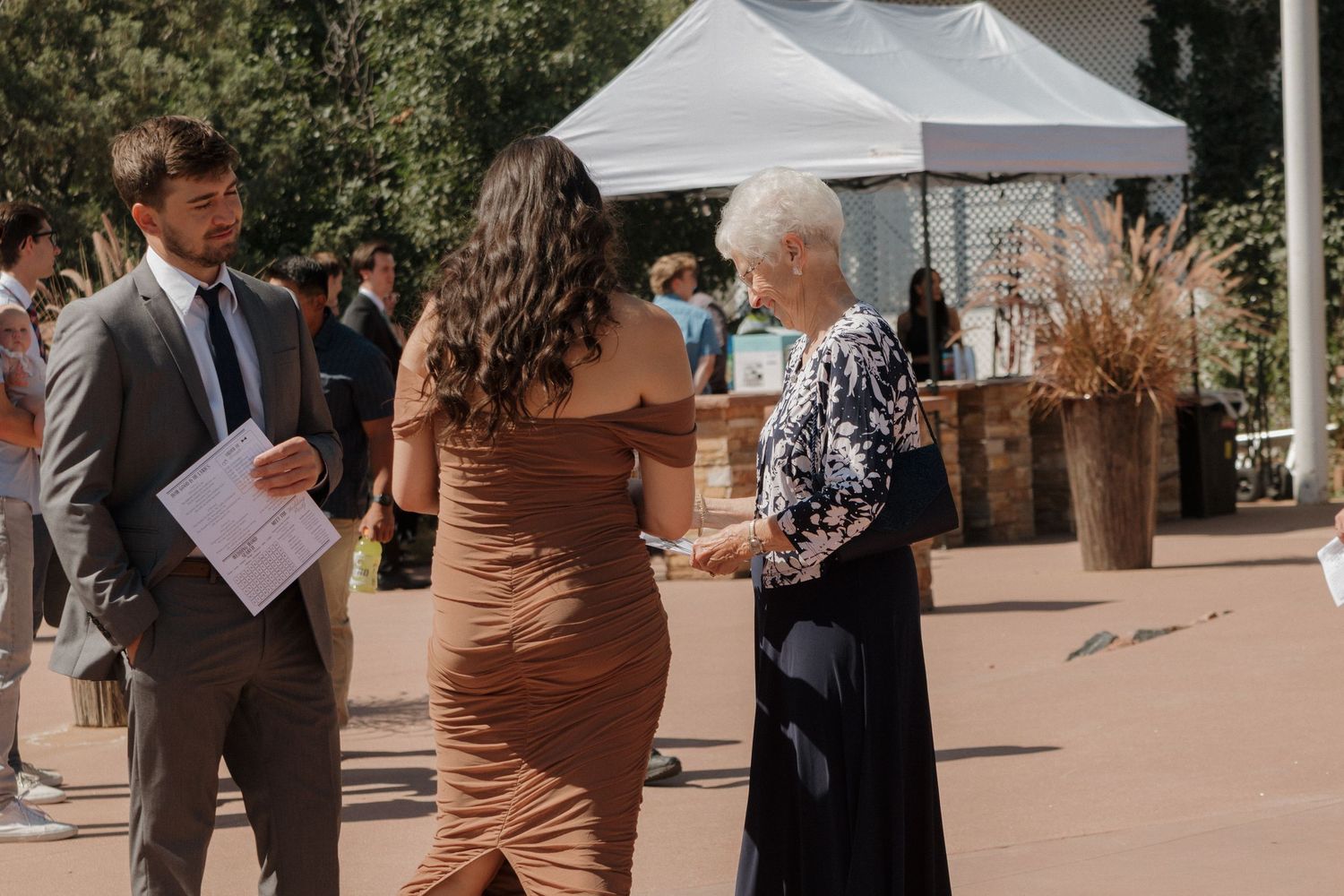 Wedding guests stand in small groups under a sunny outdoor tent during a daytime celebration.