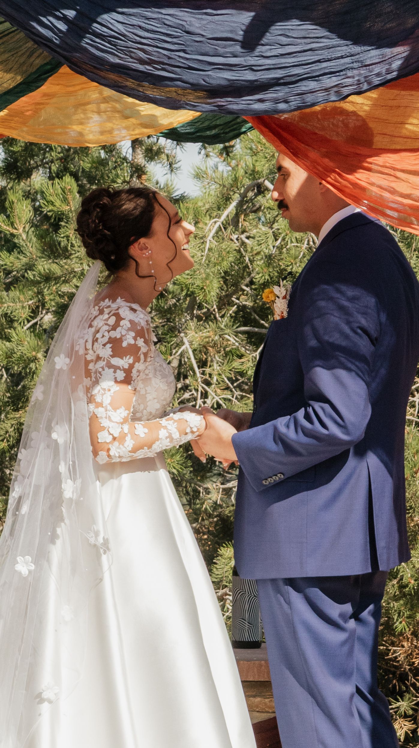 A couple in wedding attire standing under a draped fabric canopy in an outdoor garden setting.