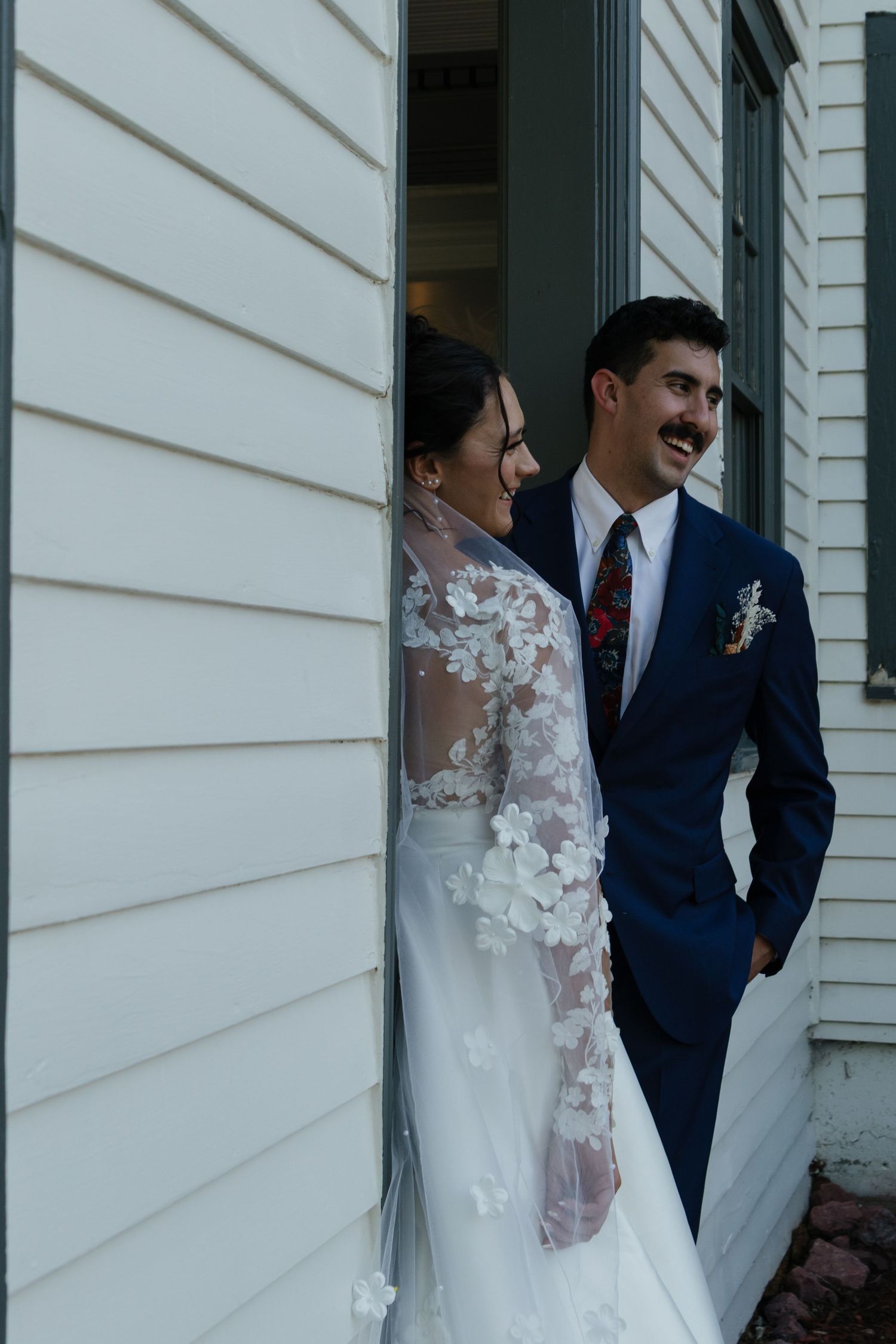 A candid moment between newlyweds along white siding of a classic building.