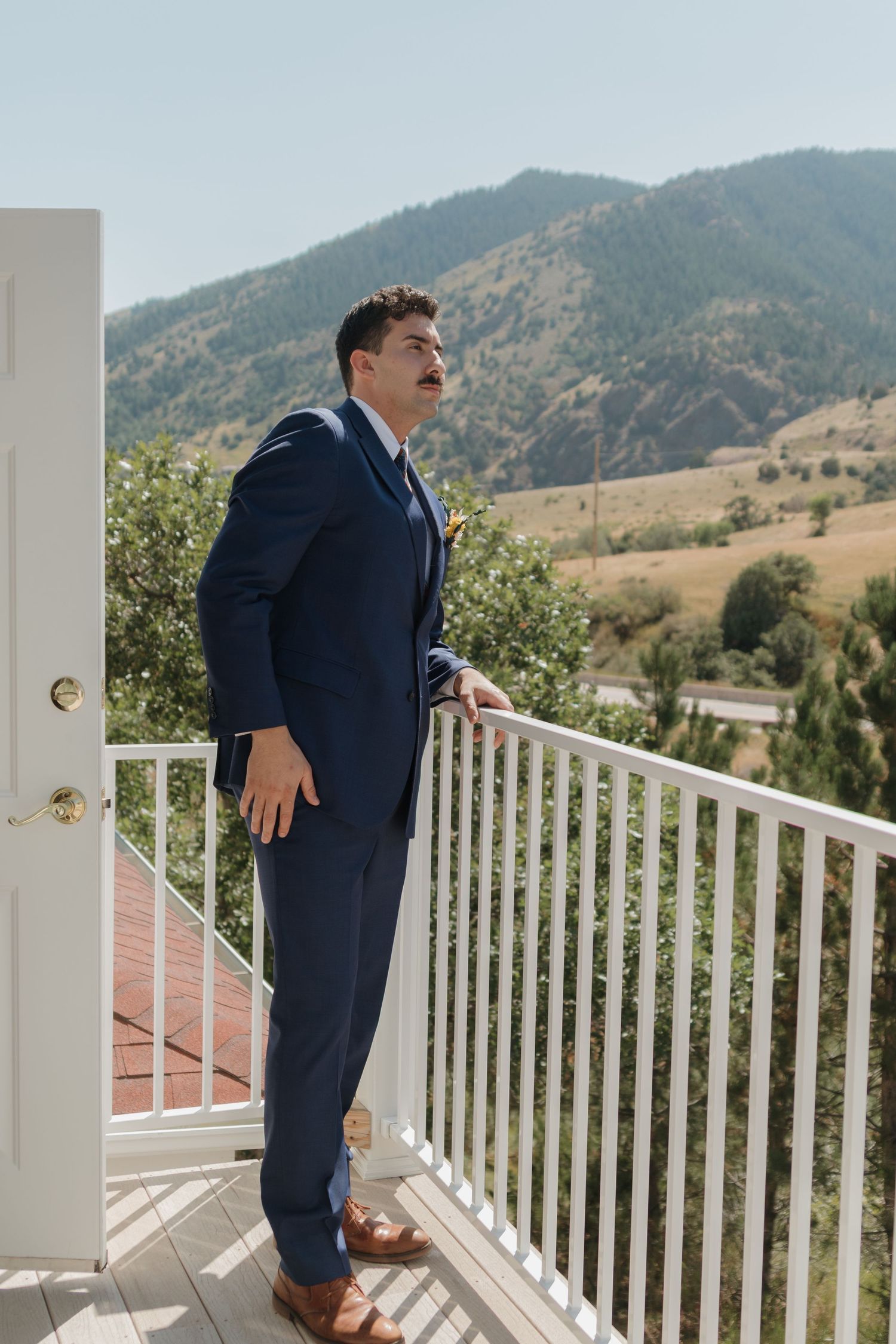 Groom in navy suit standing on white porch railing with mountain landscape backdrop.