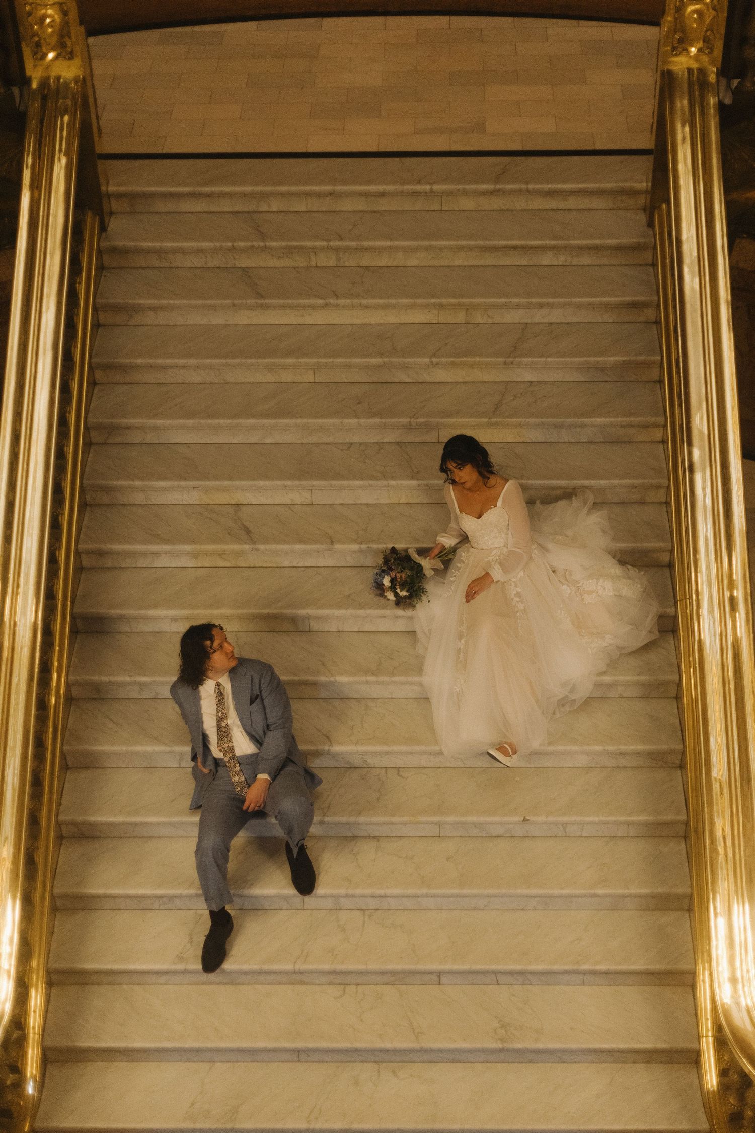 A couple poses creatively on grand marble stairs during their wedding photoshoot.