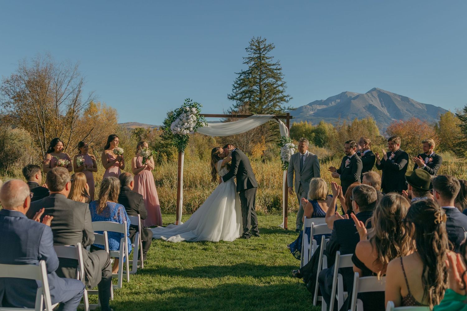Wedding ceremony guests gather outdoors with scenic mountain views and autumn trees in the background.
