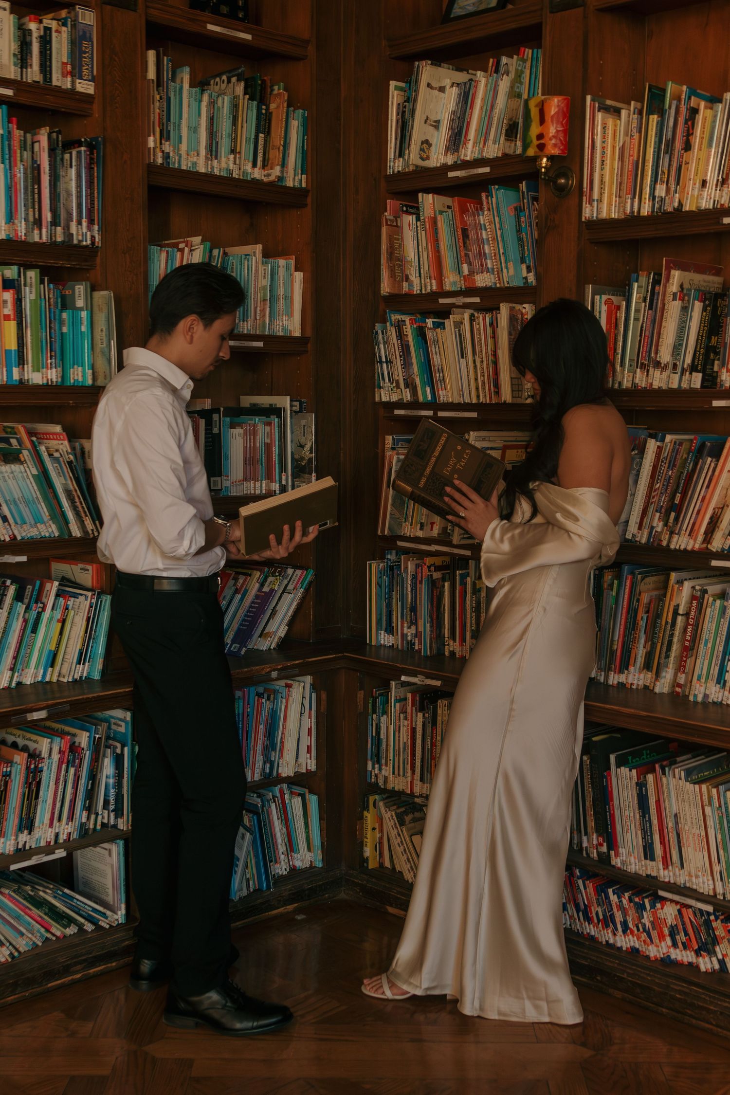 Two people browse books together in a cozy library corner filled with wooden bookshelves.
