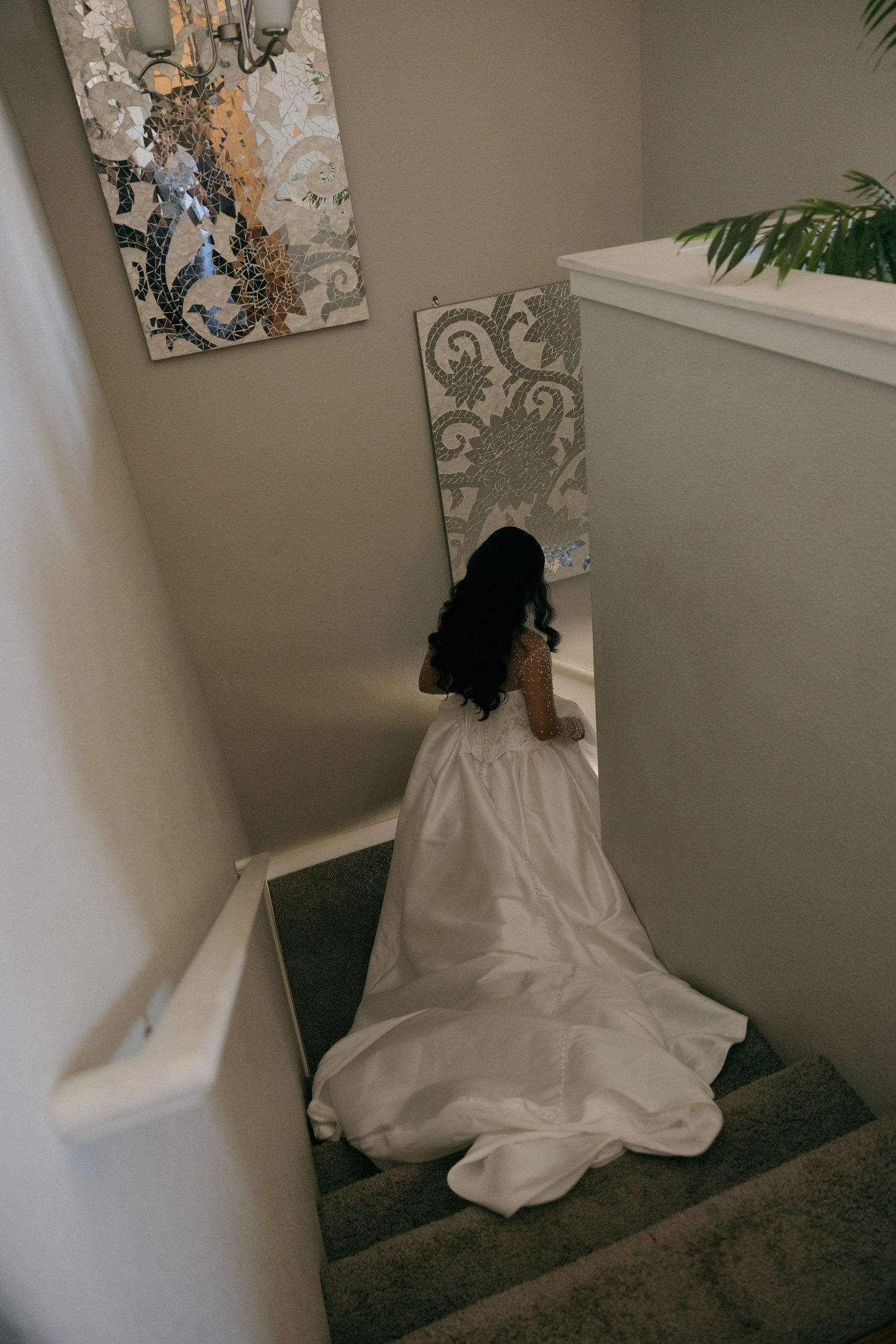 Bride in white wedding gown descending staircase viewed from above, with decorative wall art nearby.