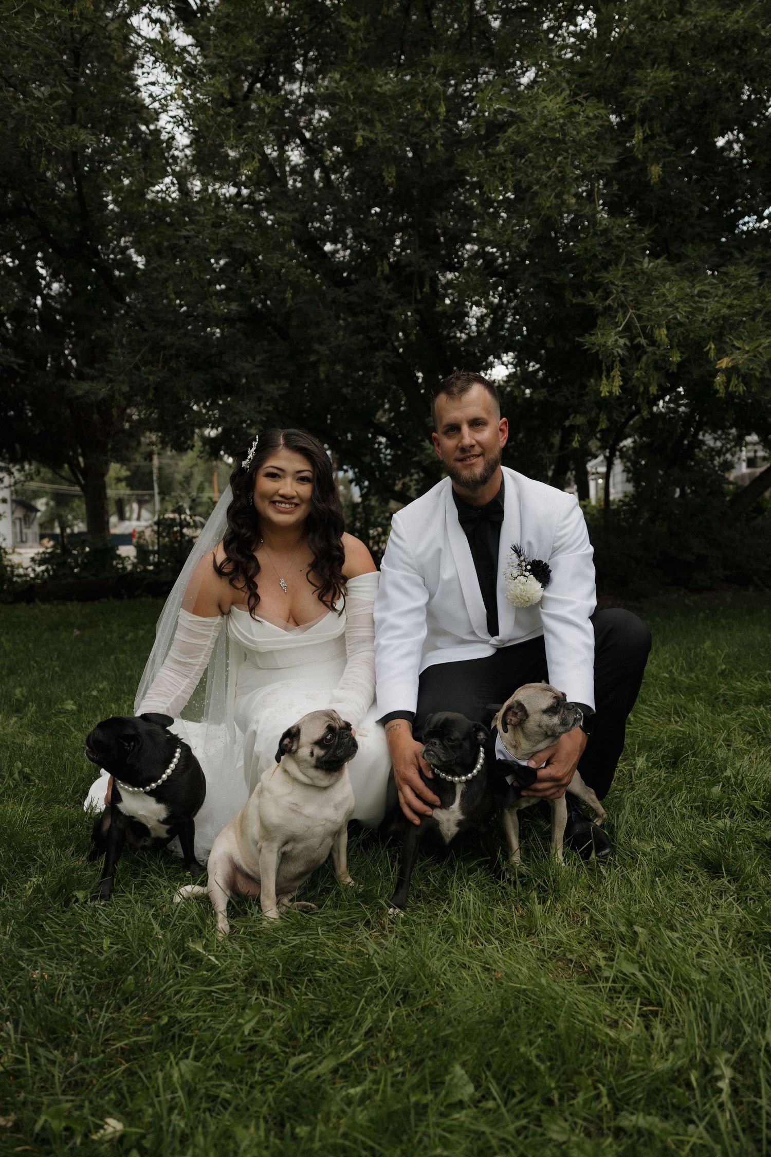 Smiling bride and groom in white tuxedo pose outdoors on grass with three pugs on wedding day.