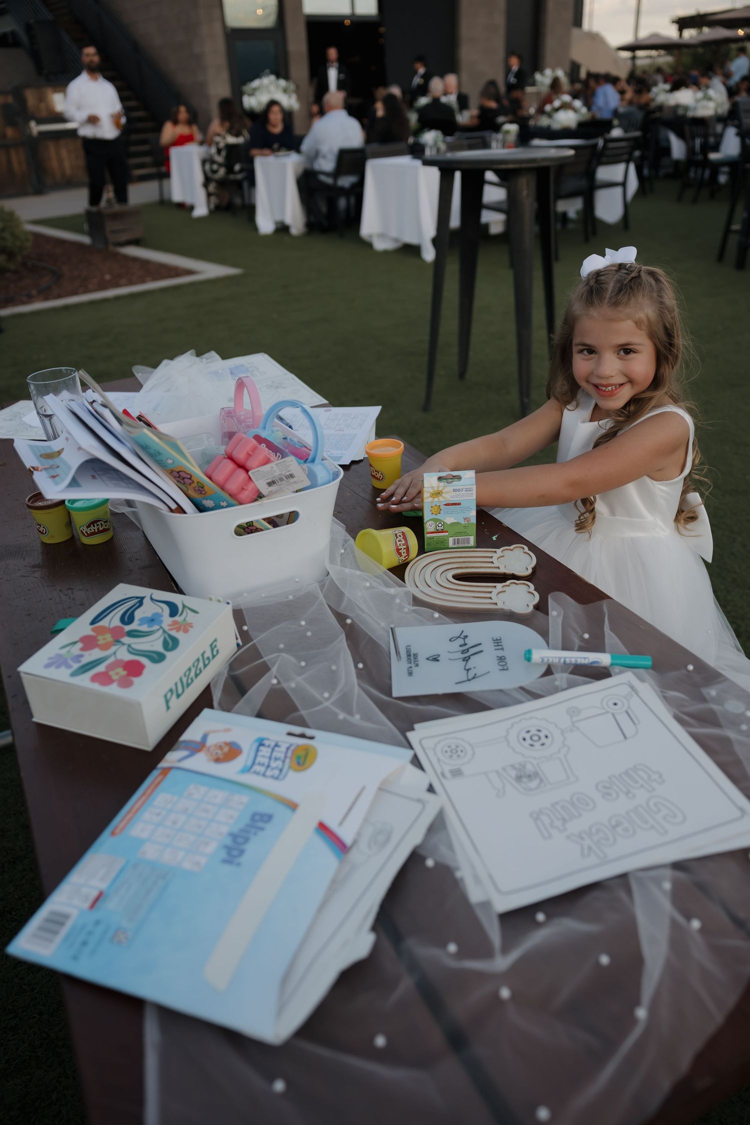 Young flower girl in white dress smiles at kids activity table during outdoor wedding reception.