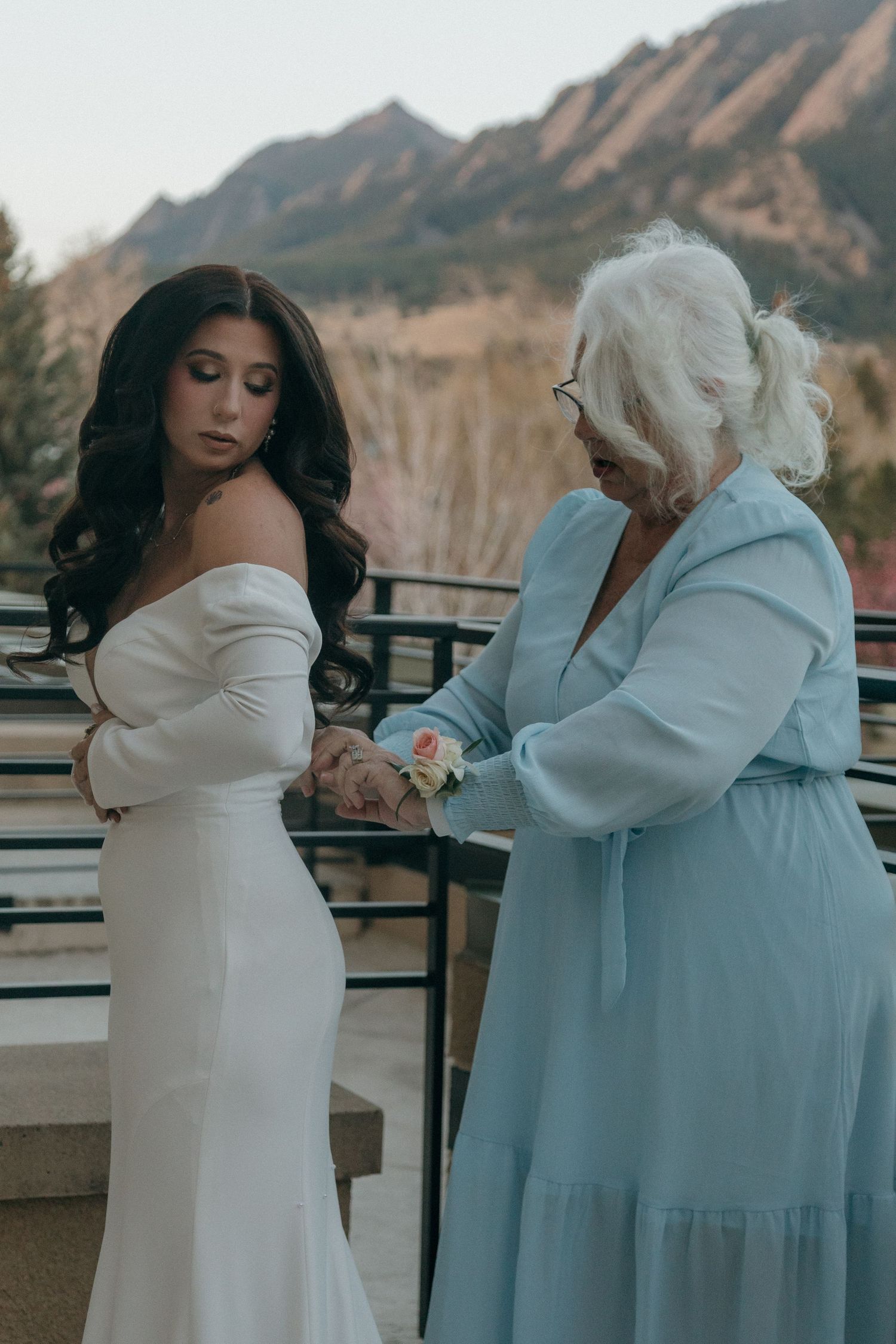 Two people in elegant dresses stand together against a mountain backdrop at sunset.