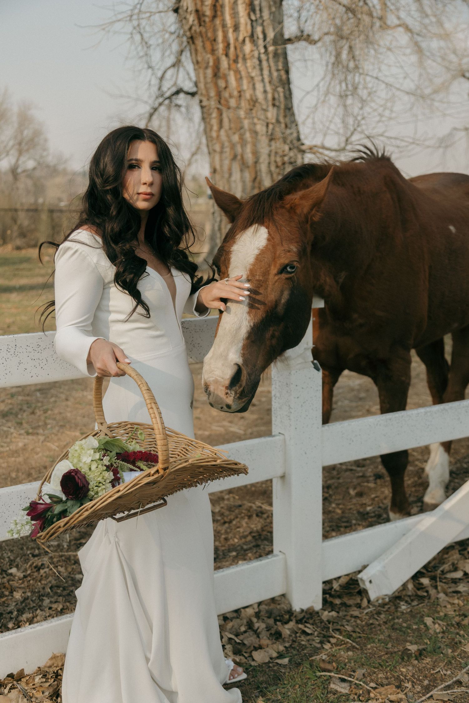 A bride in a sleek white dress stands by a white fence petting a brown horse while holding a floral basket.
